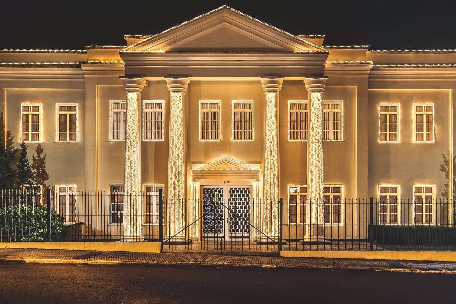 Fachada neoclássica iluminada à noite, com colunas e janelas acesas