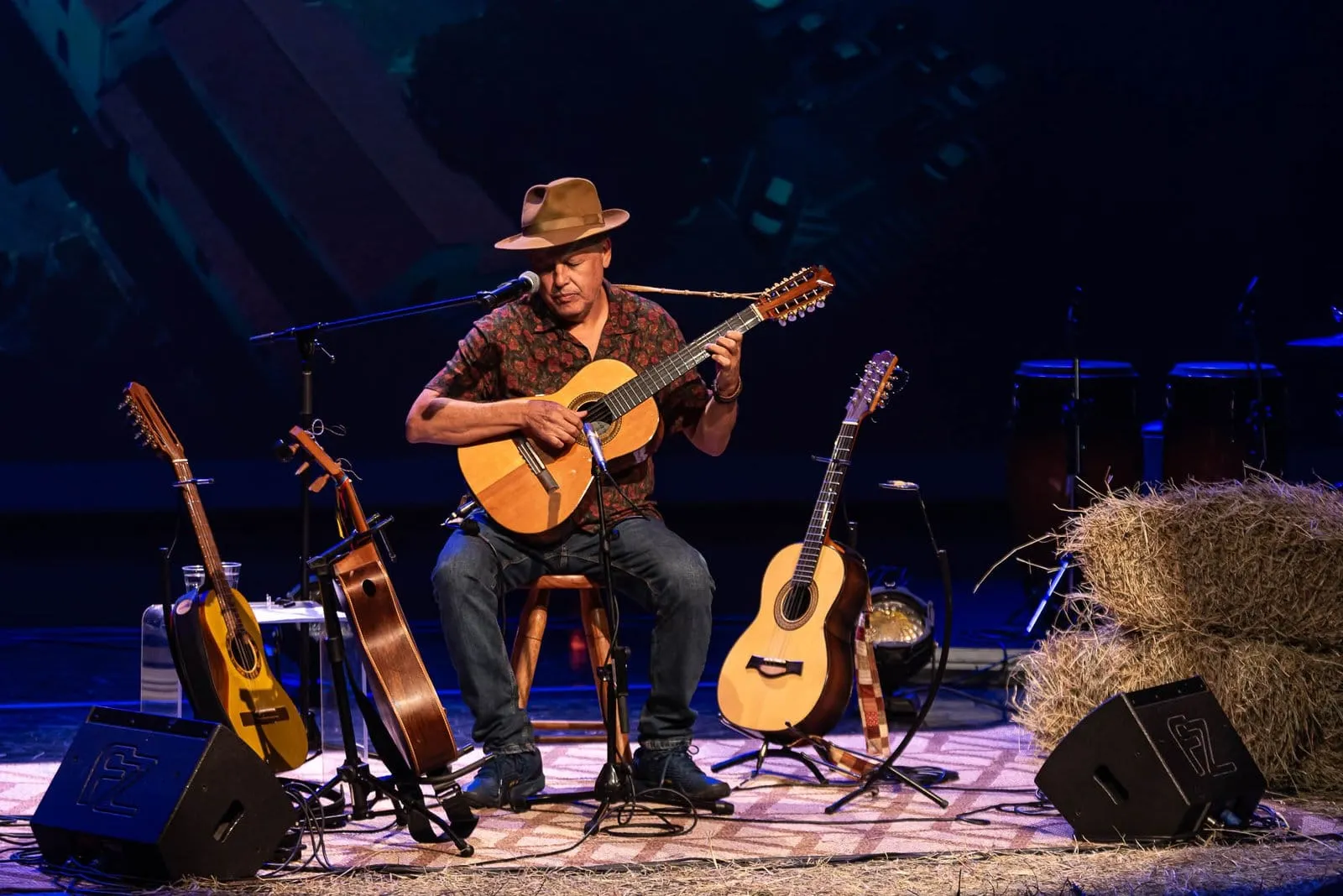 Violeiro de chapéu tocando sentado no palco, cercado de violas e fardos de feno