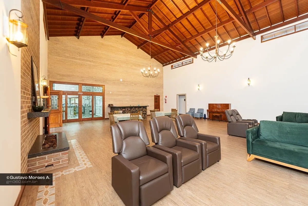 Sala de convivência com poltronas reclináveis, sofás verdes, lareira de tijolos e lustres