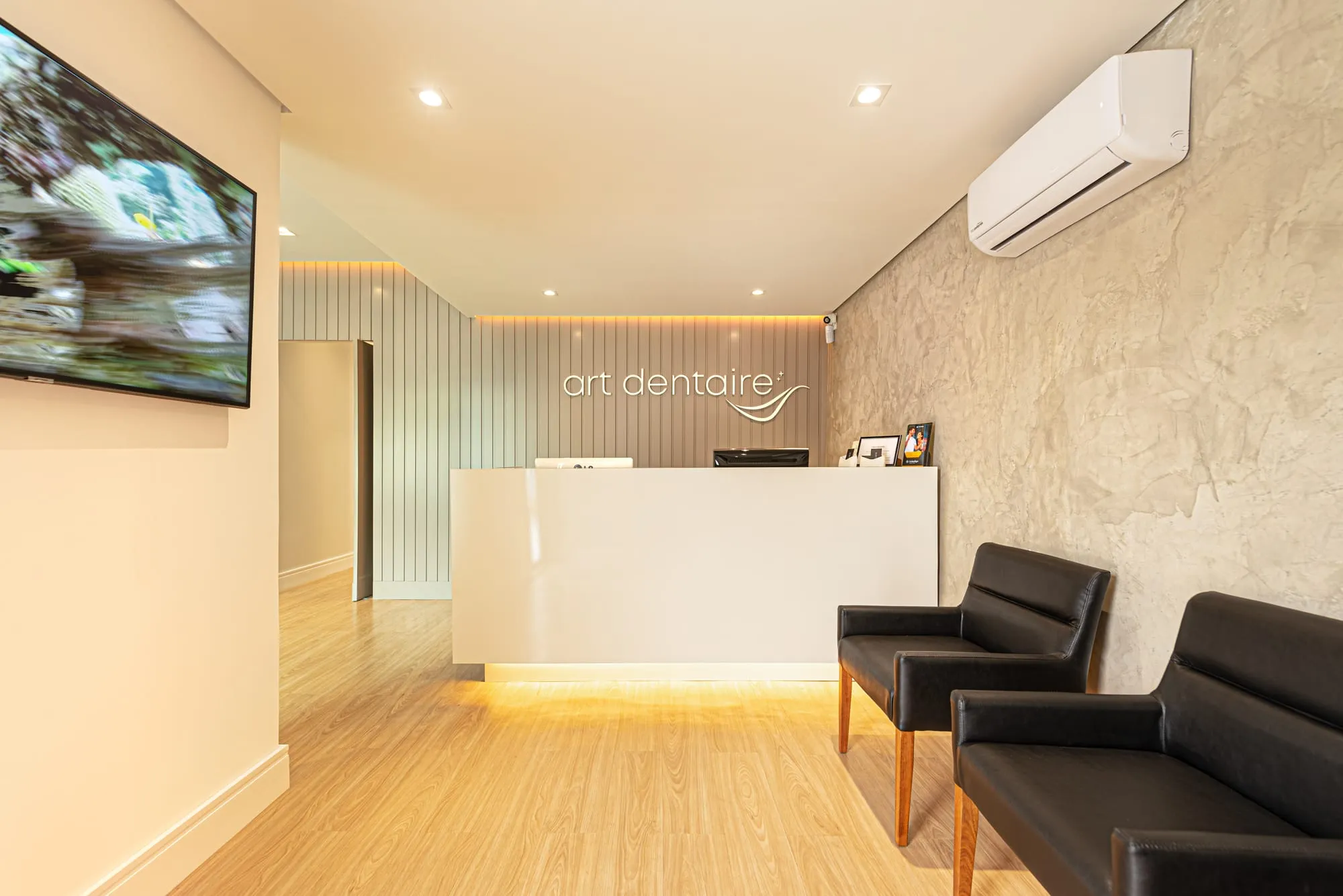 Recepção da Art Dentaire vista da entrada, com o balcão branco iluminado na base, o letreiro art dentaire na parede de ripas, a TV na parede da esquerda, duas poltronas de couro preto e piso de madeira clara