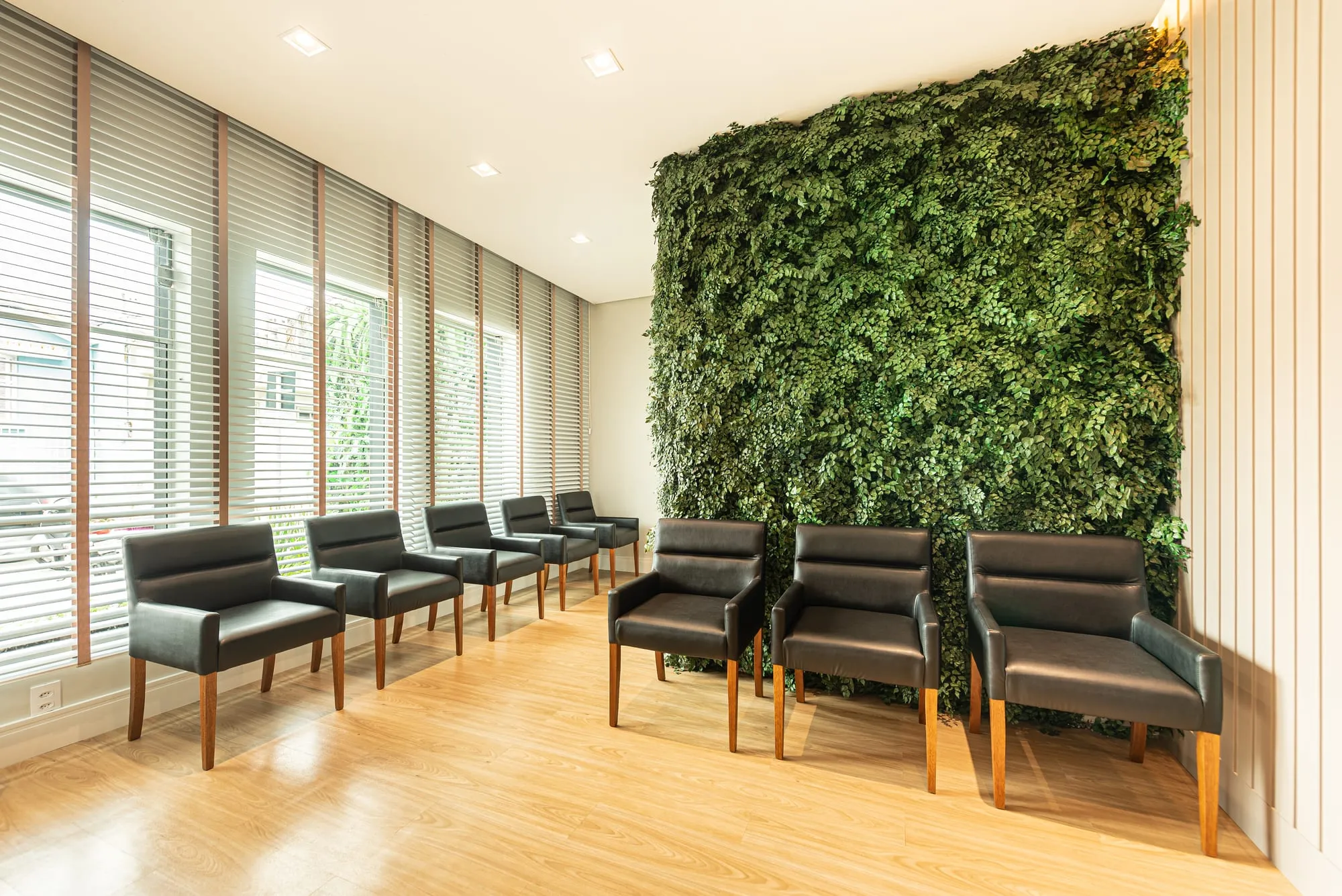 Sala de espera da Art Dentaire, com um jardim vertical de folhagem verde do chão ao teto, poltronas de couro preto ao longo das janelas com persianas de madeira, parede de ripas brancas e piso de madeira clara