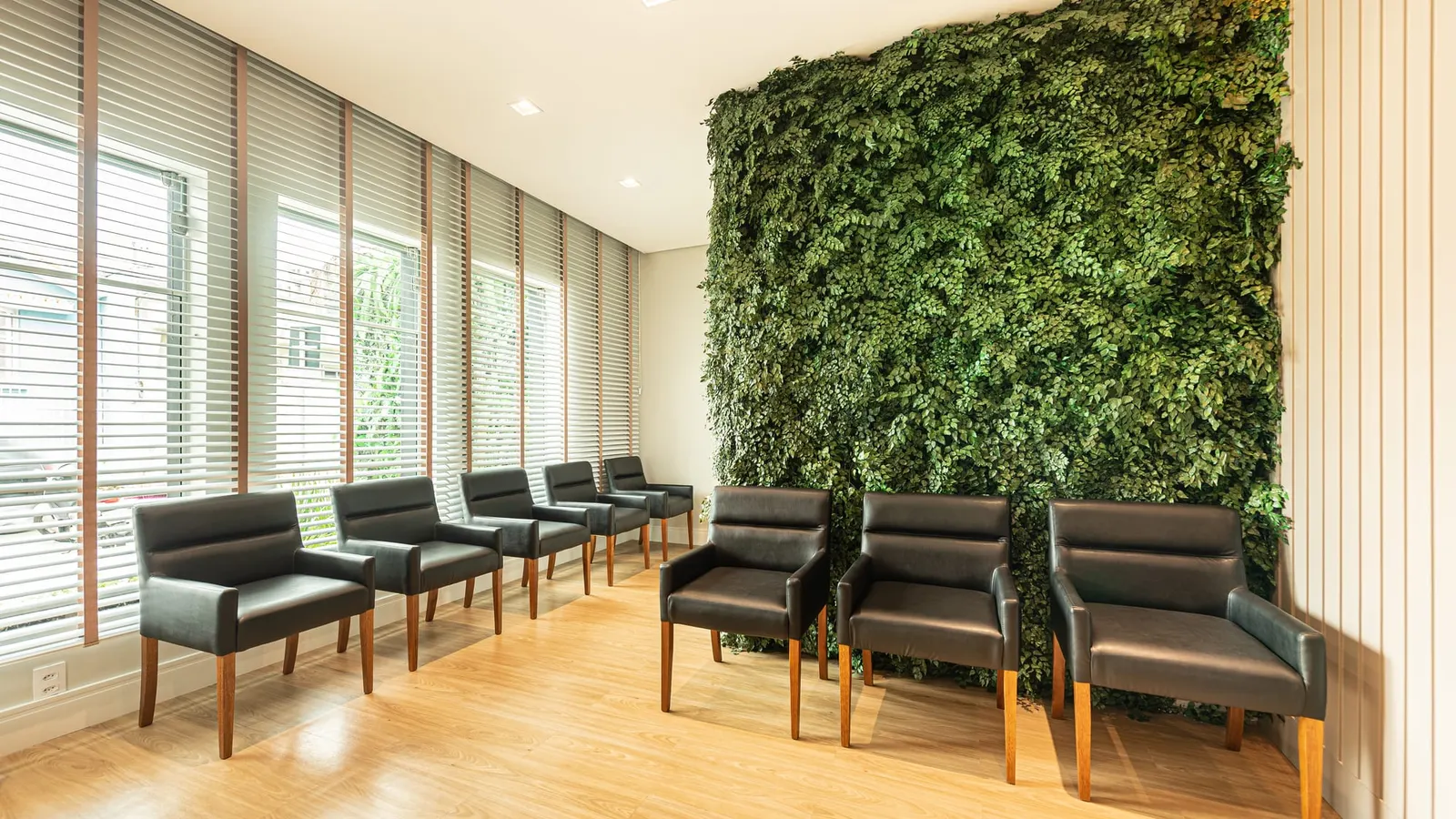 Sala de espera da Art Dentaire, com um jardim vertical de folhagem verde do chão ao teto, poltronas de couro preto ao longo das janelas com persianas de madeira, parede de ripas brancas e piso de madeira clara