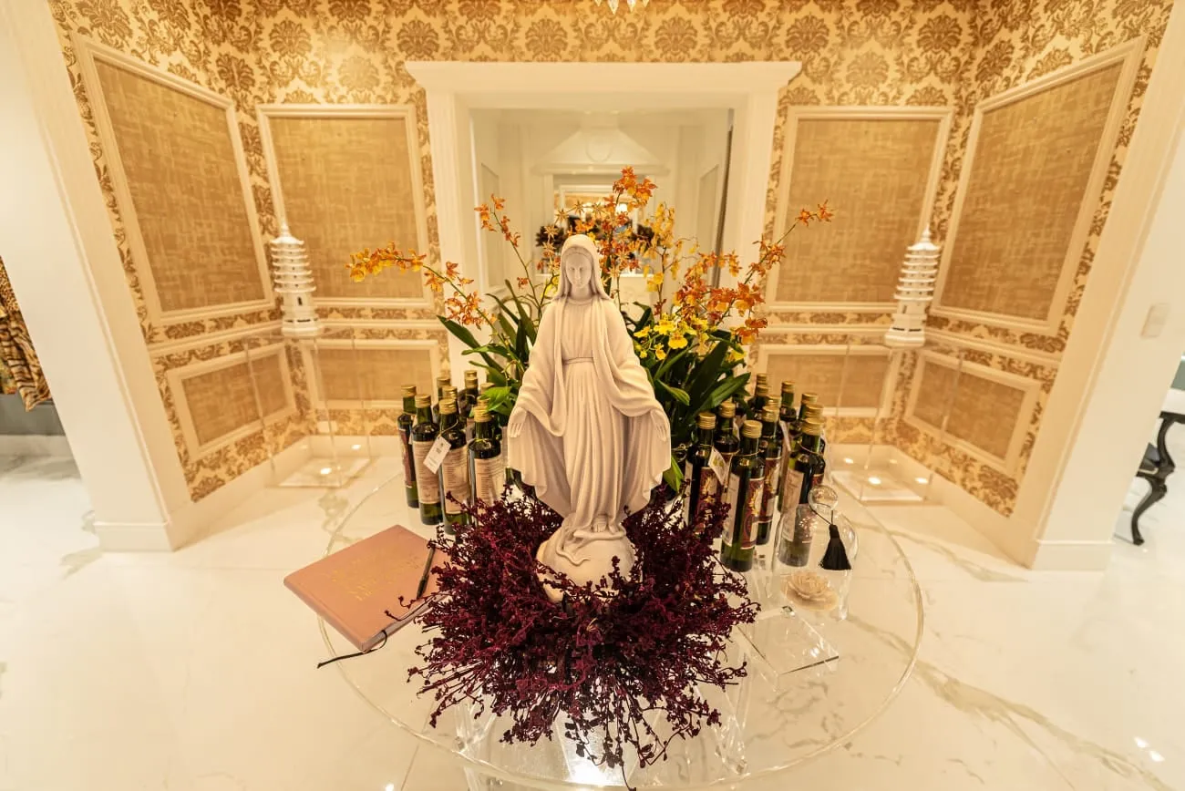 Recepção do Atelier Sandro Barros: imagem de Nossa Senhora sobre uma mesa de acrílico, entre orquídeas, garrafas e um livro, com paredes de papel adamascado dourado e piso de mármore
