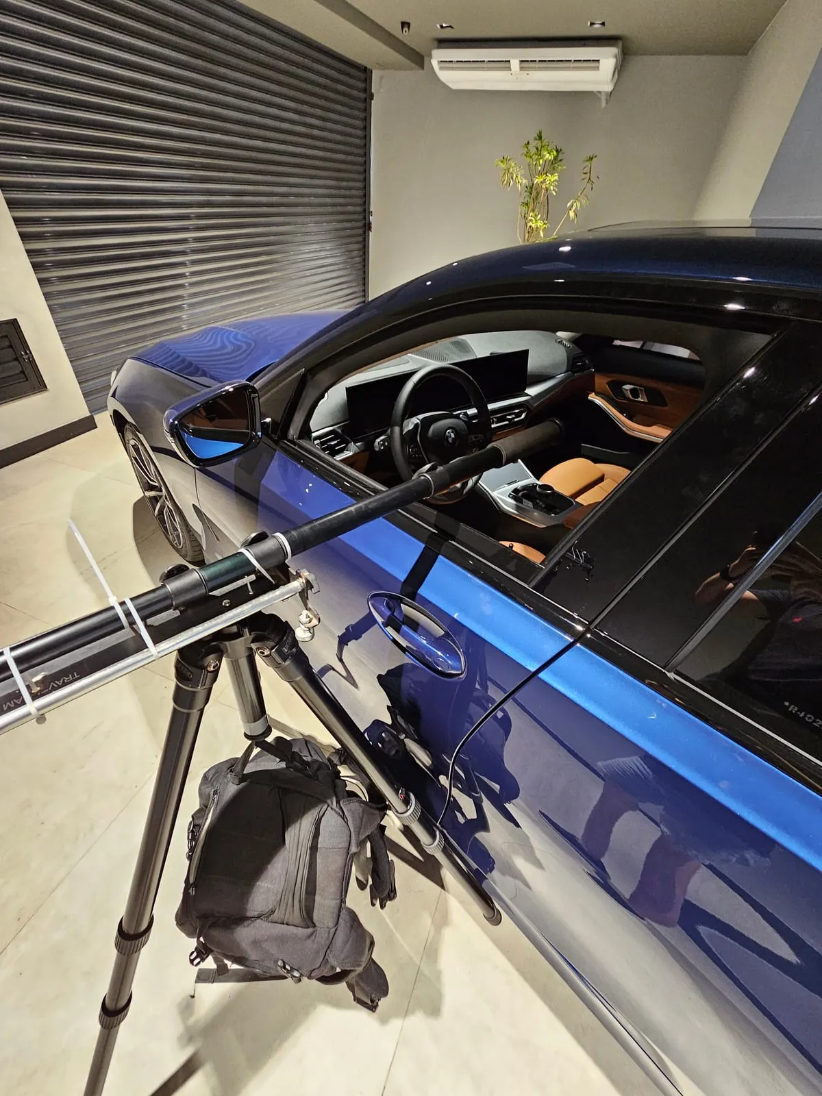 Braço extensor com ponta de espuma entrando pela janela de um sedã BMW azul, preso ao alto de um tripé, com uma mochila preta pendurada na coluna central como contrapeso e o reflexo do conjunto na porta do carro