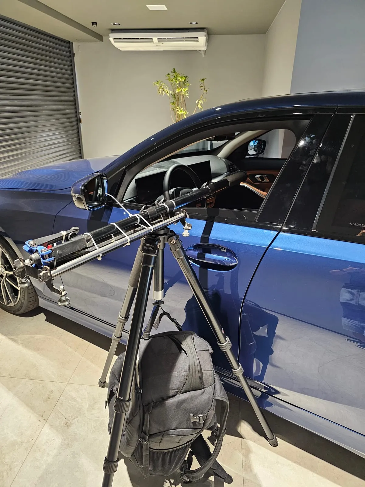 Tripé ao lado de um sedã BMW azul com um braço extensor horizontal preso por abraçadeiras brancas, entrando pela janela aberta do motorista até o meio da cabine, com bancos de couro caramelo e painel de telas visíveis por dentro e uma mochila preta pendurada embaixo do tripé