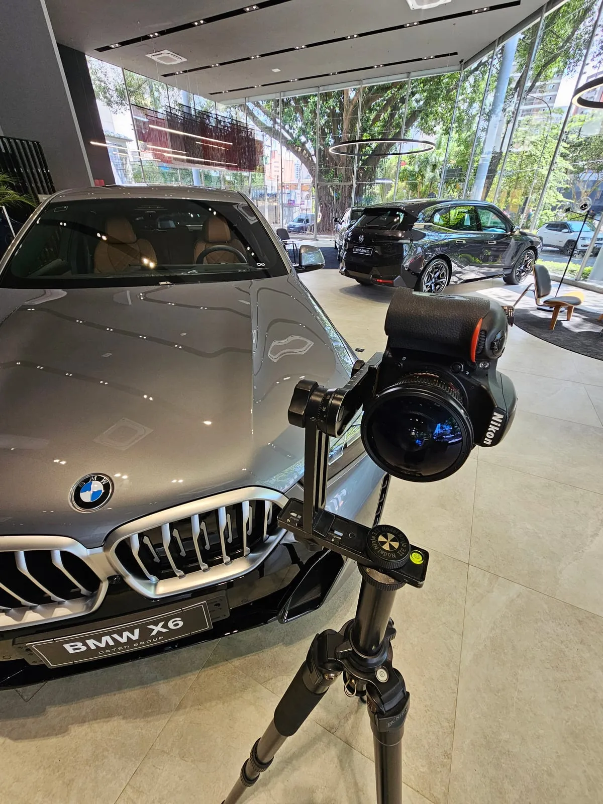 Câmera Nikon com lente olho de peixe montada numa cabeça panorâmica sobre um tripé, de frente para a grade de um BMW X6 prata com a placa de exposição BMW X6 Osten Group, no salão envidraçado da BMW Osten Perdizes, com um BMW iX escuro e as árvores da avenida ao fundo
