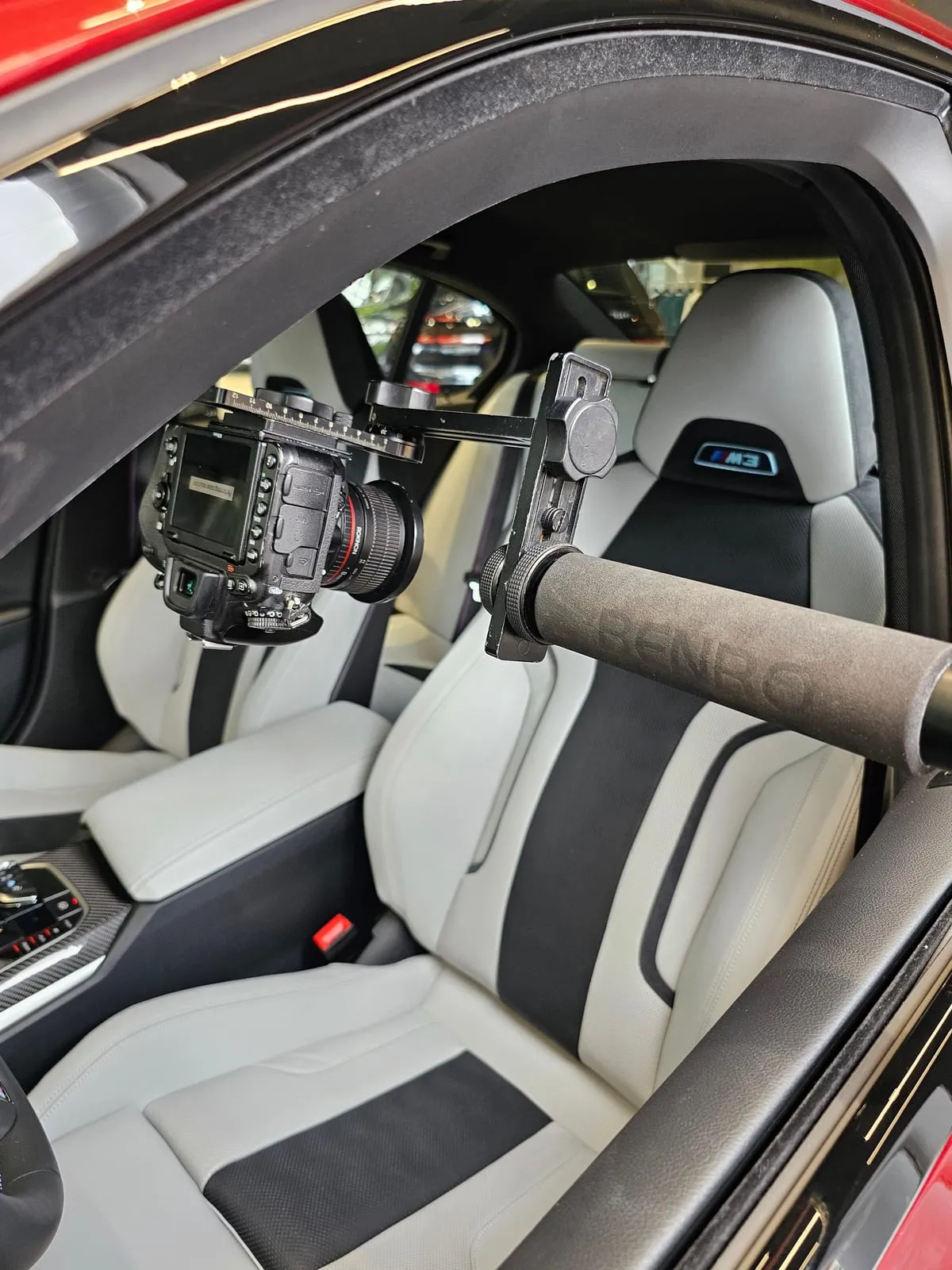 Câmera presa a uma cabeça panorâmica na ponta do braço extensor, com espuma no cabo, dentro de um BMW M vermelho, entre os bancos esportivos claros com faixas pretas e o logo M3 no encosto