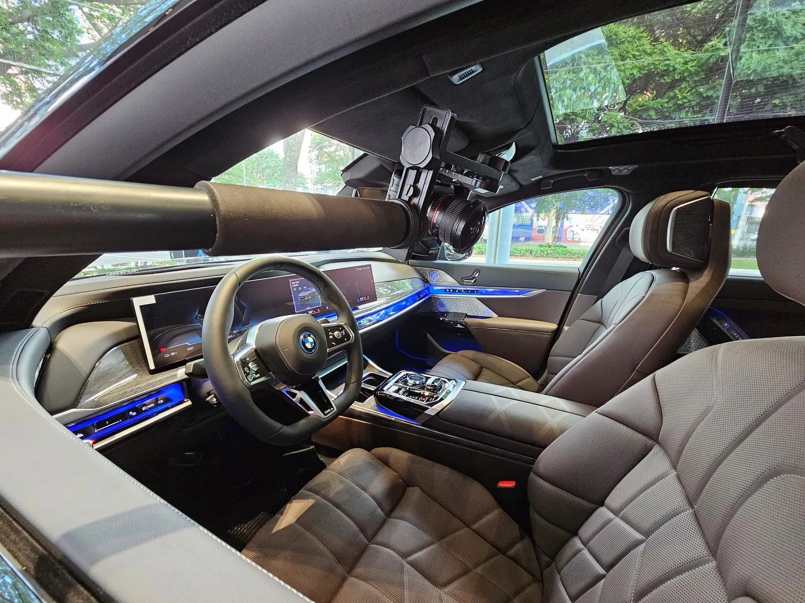 Câmera com lente olho de peixe presa a uma cabeça panorâmica na ponta de um braço extensor, no centro da cabine de um BMW i7 vazio, com o volante, o painel curvo, a faixa de luz azul nas portas, os bancos de couro e o teto de vidro com as árvores da avenida acima