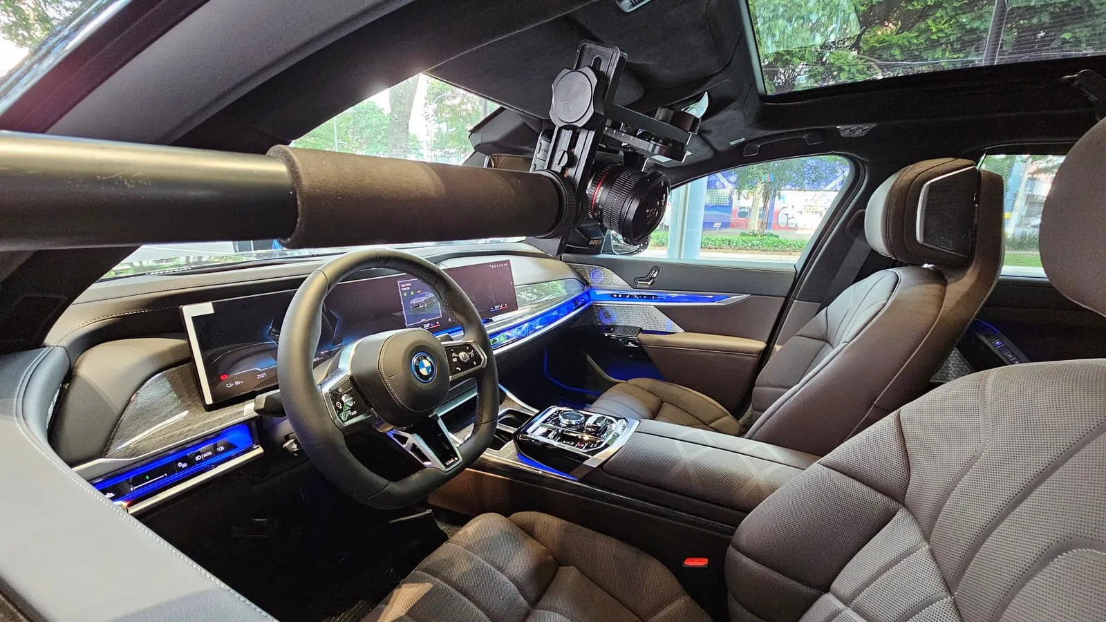 Câmera com lente olho de peixe presa a uma cabeça panorâmica na ponta de um braço extensor, no centro da cabine de um BMW i7 vazio, com o volante, o painel curvo, a faixa de luz azul nas portas, os bancos de couro e o teto de vidro com as árvores da avenida acima