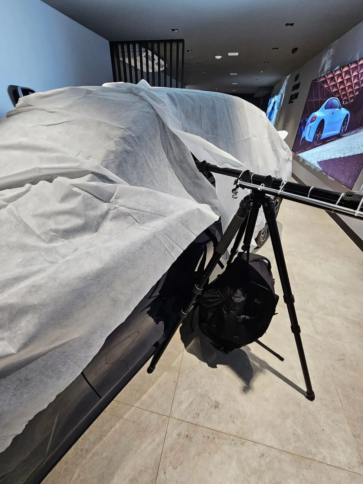 Detalhe do tripé encostado na lateral de um carro coberto por manta branca, com o braço extensor preso por abraçadeiras e a mochila preta pendurada embaixo