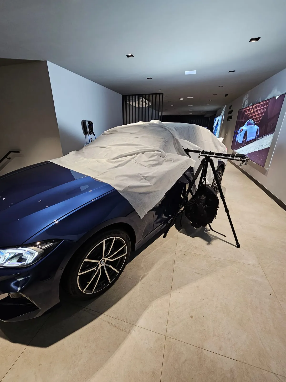 Sedã BMW azul com o teto e os vidros cobertos por uma manta branca, com o tripé e o braço extensor ao lado da porta e uma mochila pendurada, num corredor da loja com telões na parede