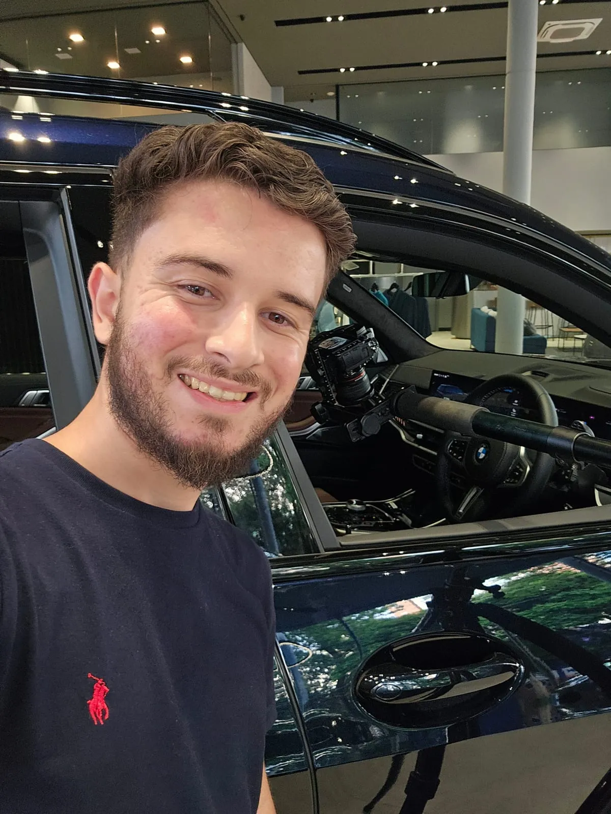 Gustavo Moraes Augusto, fundador da gm360, sorrindo numa selfie ao lado de um BMW X7 azul-escuro, com a câmera presa ao braço extensor dentro da cabine, perto do volante, no salão da BMW Osten Perdizes