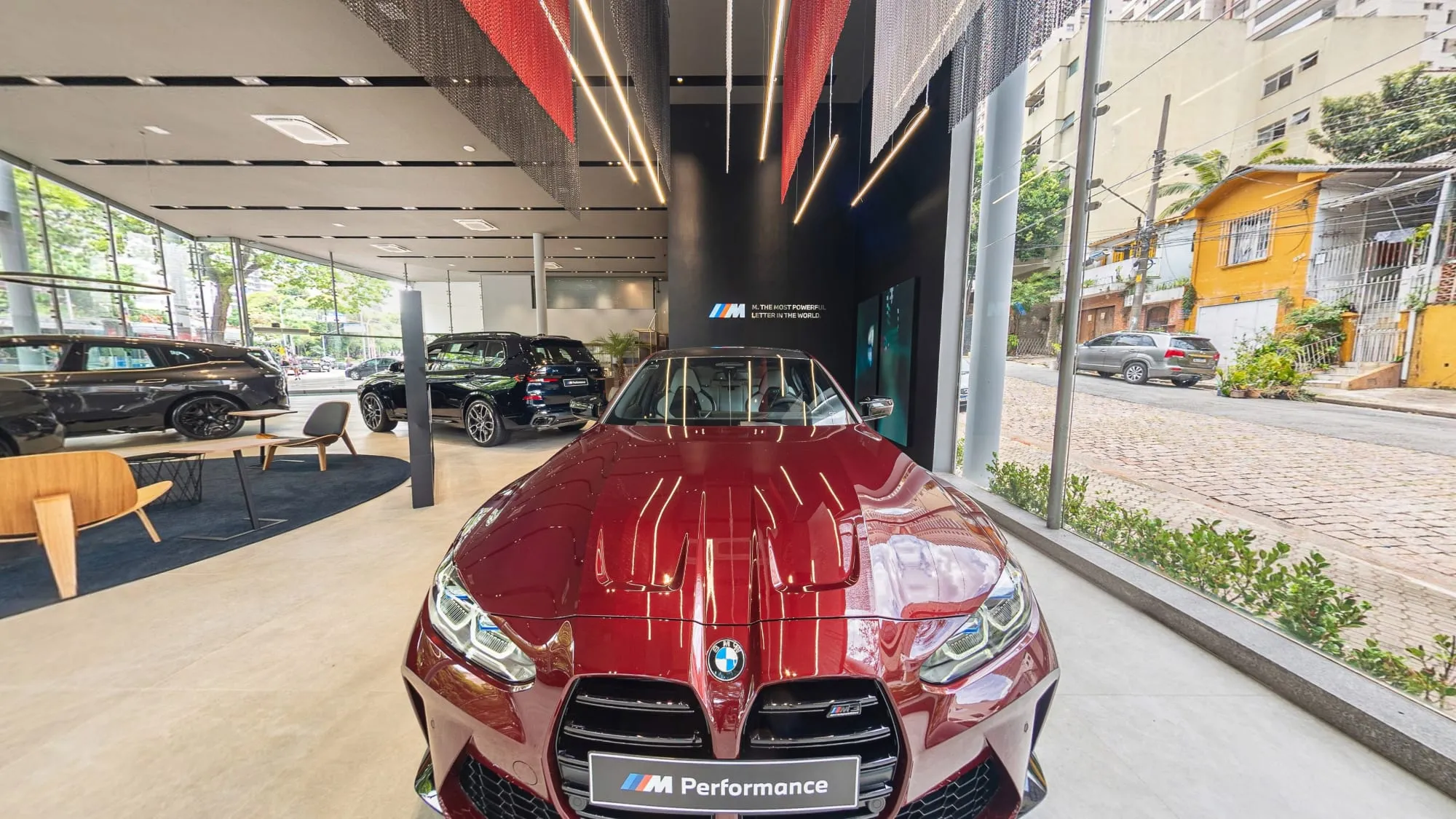 Panorâmica da Área M da BMW Osten Perdizes, com um BMW M vermelho de frente, com placa M Performance, diante de uma parede escura com o logo M, outros BMW expostos ao lado e a vitrine para a rua
