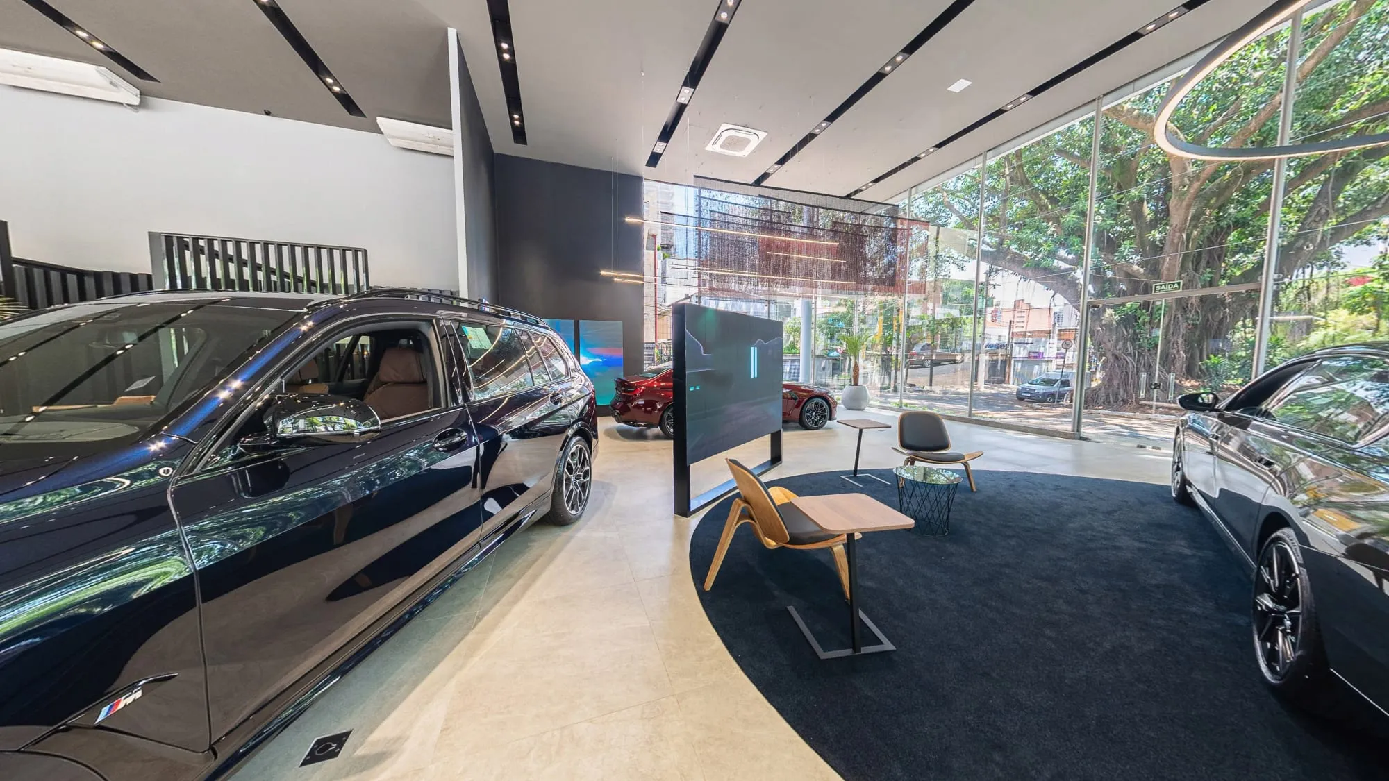 Panorâmica do espaço dos elétricos da BMW Osten Perdizes, com um SUV BMW azul-escuro à esquerda, um sedã escuro à direita, um totem de tela e poltronas de madeira sobre tapete azul, diante da vitrine de vidro para a avenida arborizada