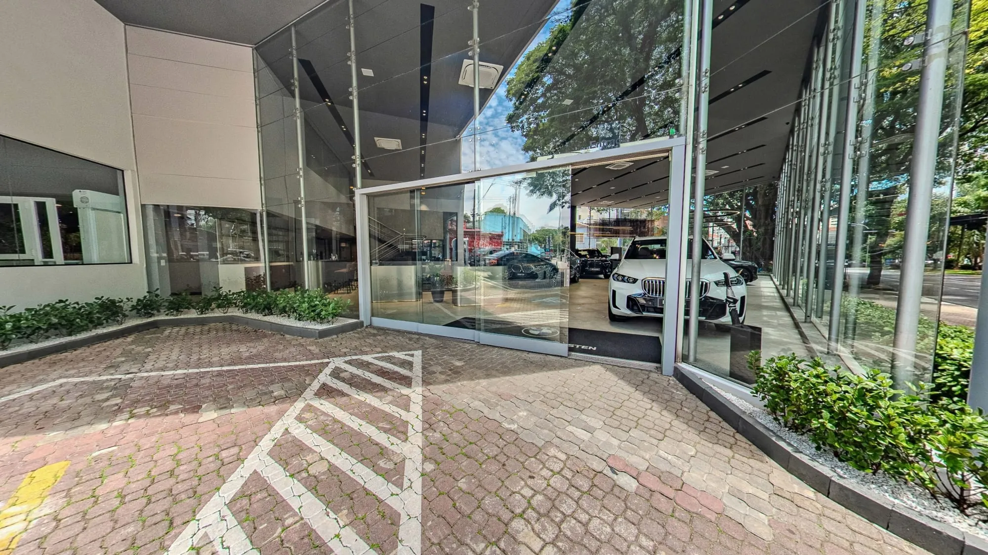 Panorâmica da entrada da BMW Osten Perdizes, com o piso de blocos intertravados do estacionamento, a faixa de pedestres pintada, a porta de vidro aberta, um BMW X5 branco visível no salão e canteiros de plantas ao longo da fachada envidraçada