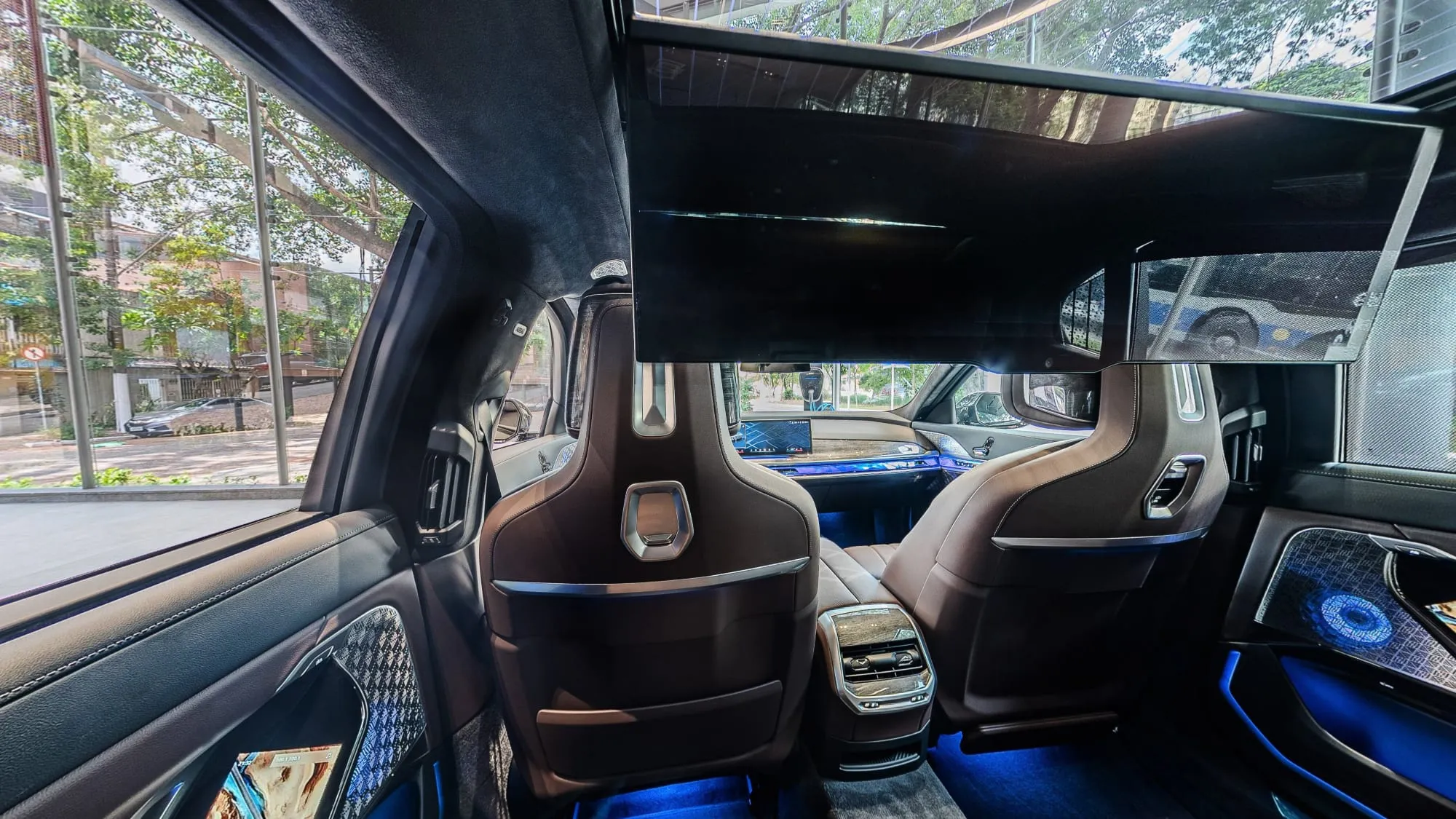 Panorâmica do banco de trás do BMW i7, com a tela de cinema baixada do teto à frente, os encostos dos bancos dianteiros em couro marrom, o console traseiro, as telas nas portas, a luz azul no assoalho e a vitrine da loja pela janela