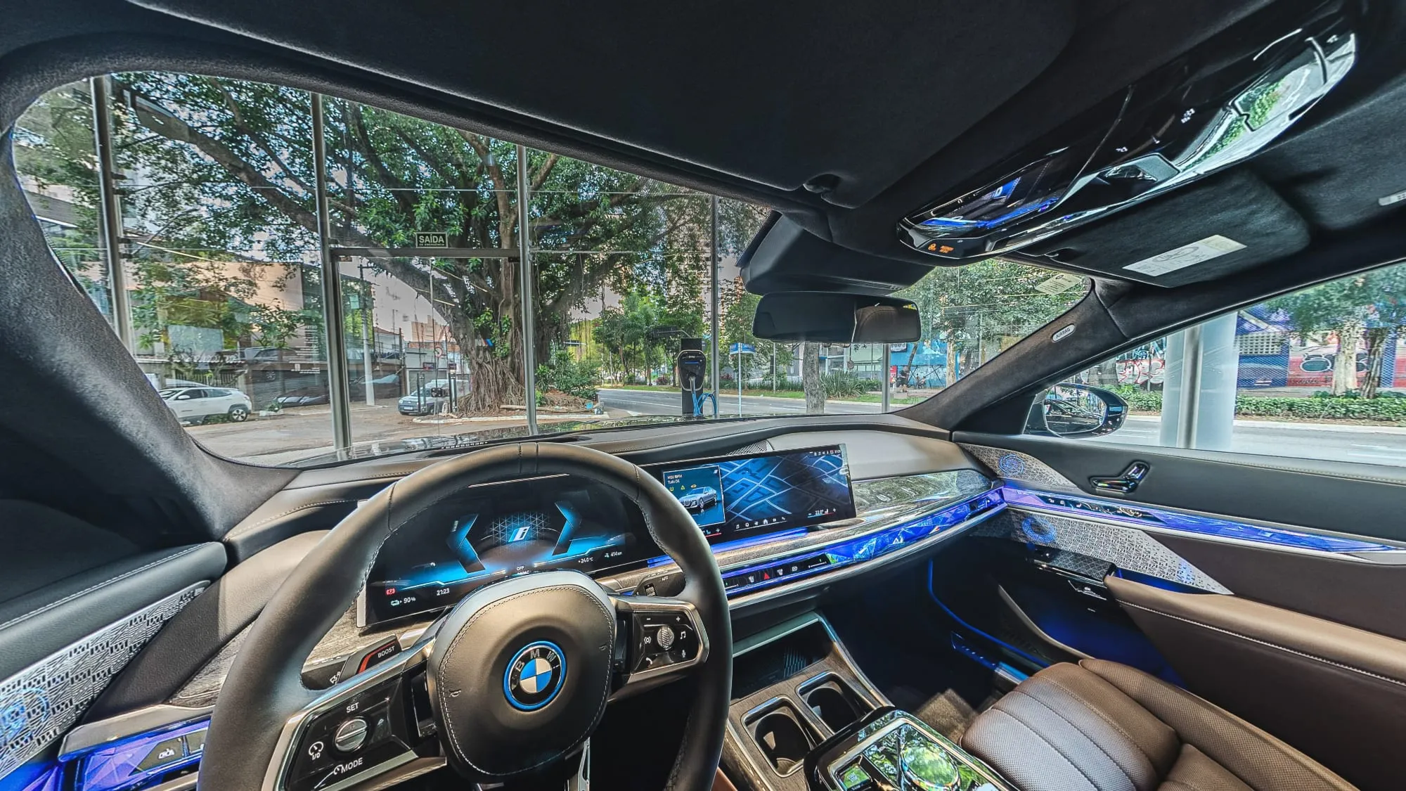 Panorâmica do interior do BMW i7 vista do banco do motorista, com o volante, o painel curvo de telas, a faixa de luz azul no painel e na porta, bancos de couro marrom e, pelo para-brisa, a vitrine da loja, o carregador elétrico na calçada e as árvores da avenida