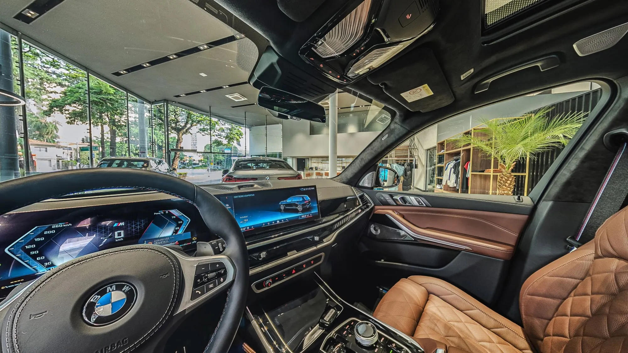 Panorâmica do interior do BMW X7 vista do banco do motorista, com o volante, o painel de telas, bancos de couro caramelo matelassê e, pelos vidros, o salão da BMW Osten Perdizes com outro BMW, a área de roupas e uma palmeira