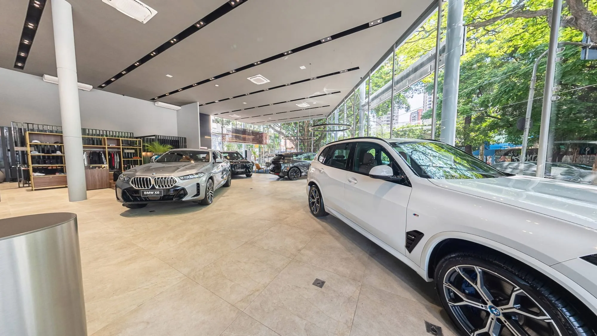 Panorâmica do salão da BMW Osten Perdizes, em São Paulo, com um BMW X5 branco em primeiro plano, um X6 prata e outros modelos ao fundo, a área de roupas e acessórios à esquerda e a fachada de vidro com as árvores da avenida