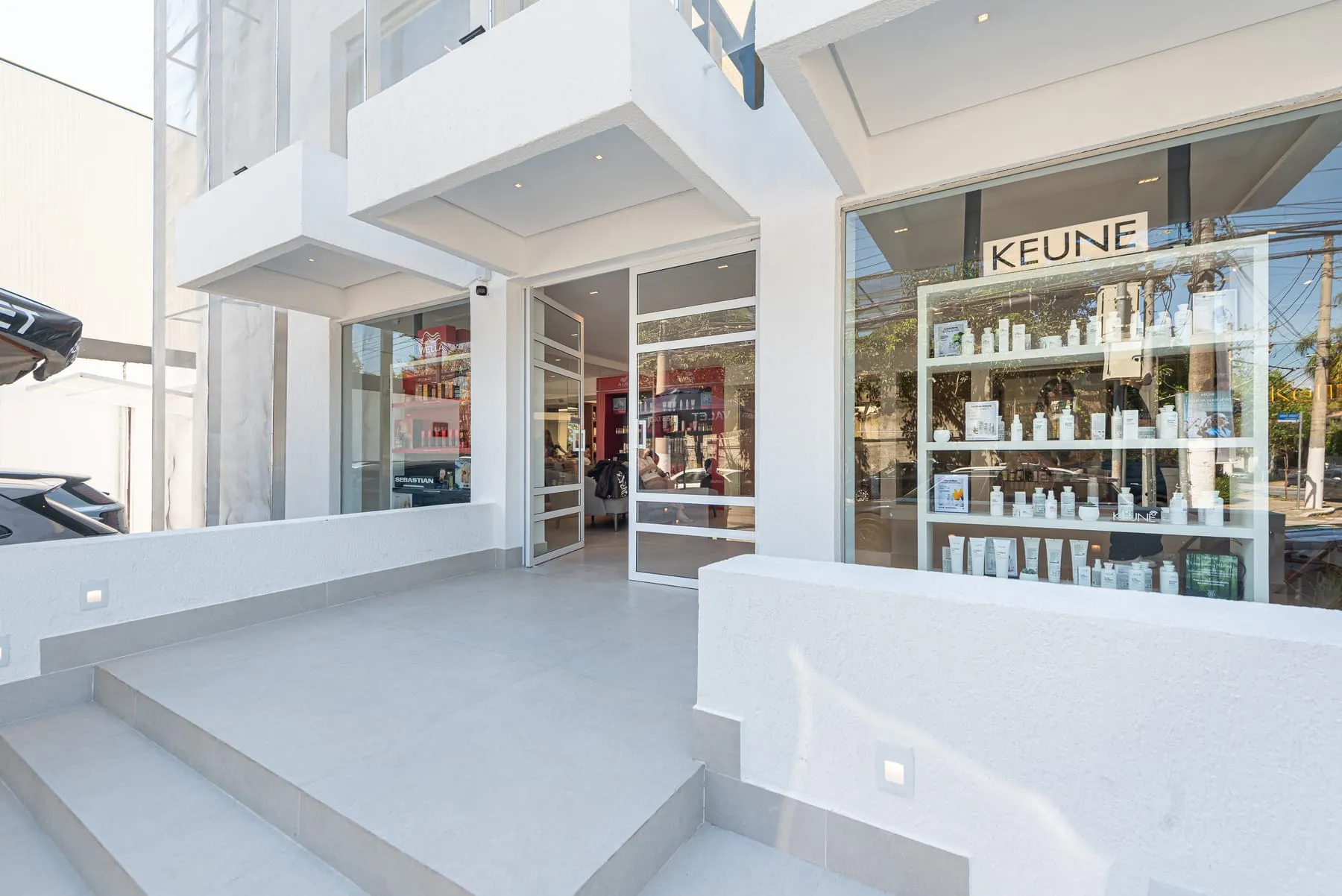 Degraus de porcelanato claro levam à porta de vidro da entrada, sob volumes brancos em balanço, com a vitrine Keune iluminada à direita