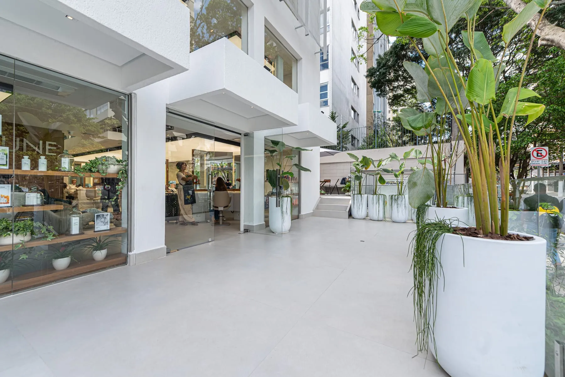 Terraço externo ao lado da vitrine Keune, com piso claro, vasos brancos altos com plantas de folhas grandes e a porta de vidro que dá para o salão