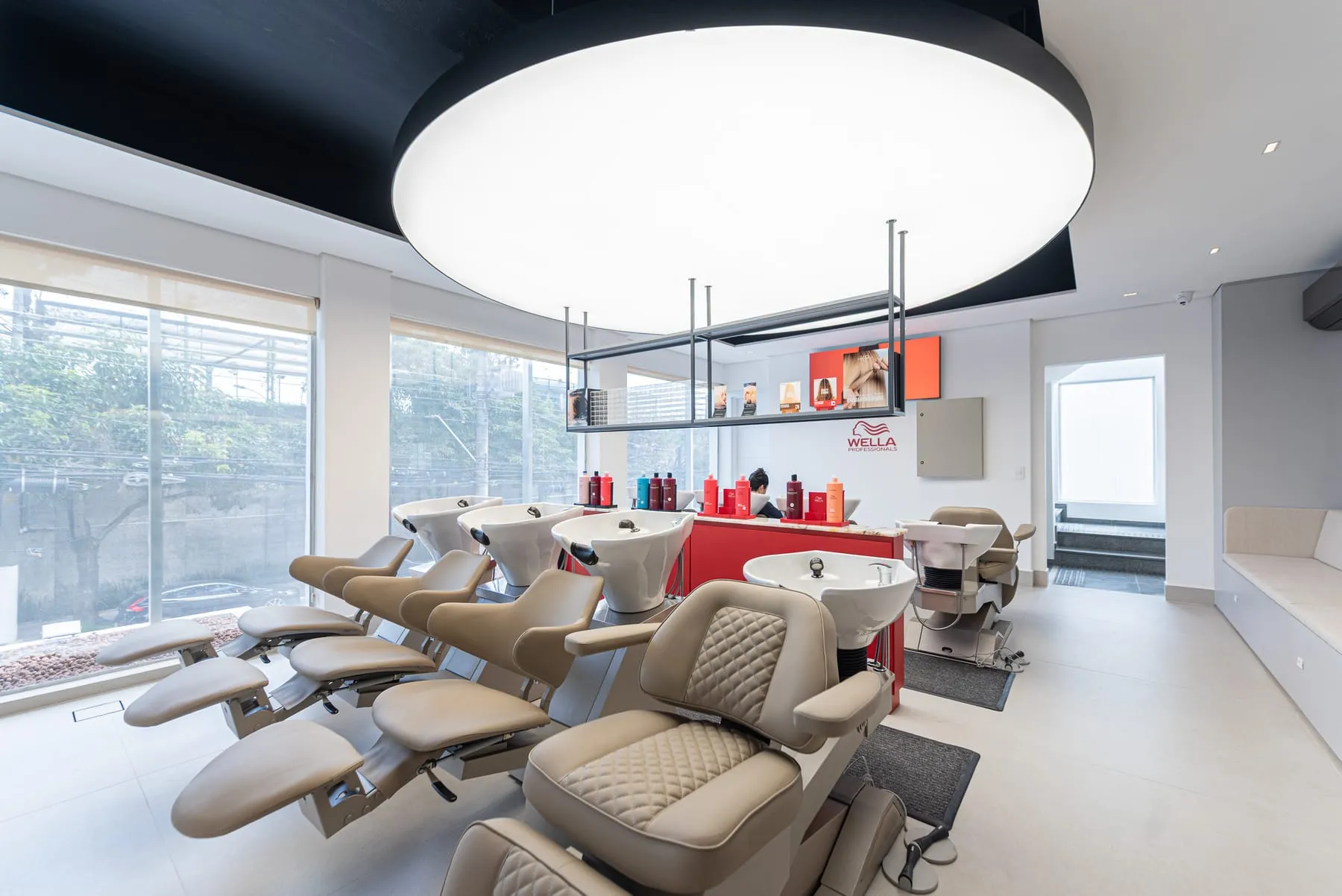 Lavatórios Wella no 1º piso do C.Kamura, com poltronas bege em capitonê, ilha vermelha com produtos, prateleira metálica suspensa, painel de LED e luminária circular no teto preto