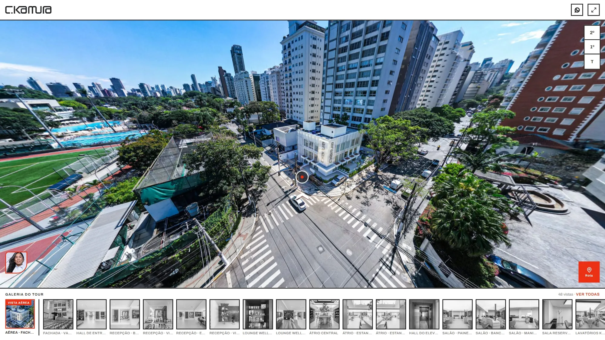 Vista aérea de drone dentro do tour, com o prédio branco de esquina do C.Kamura no centro, a faixa de pedestres, árvores e prédios altos em volta e o ponto de navegação na calçada que leva à fachada