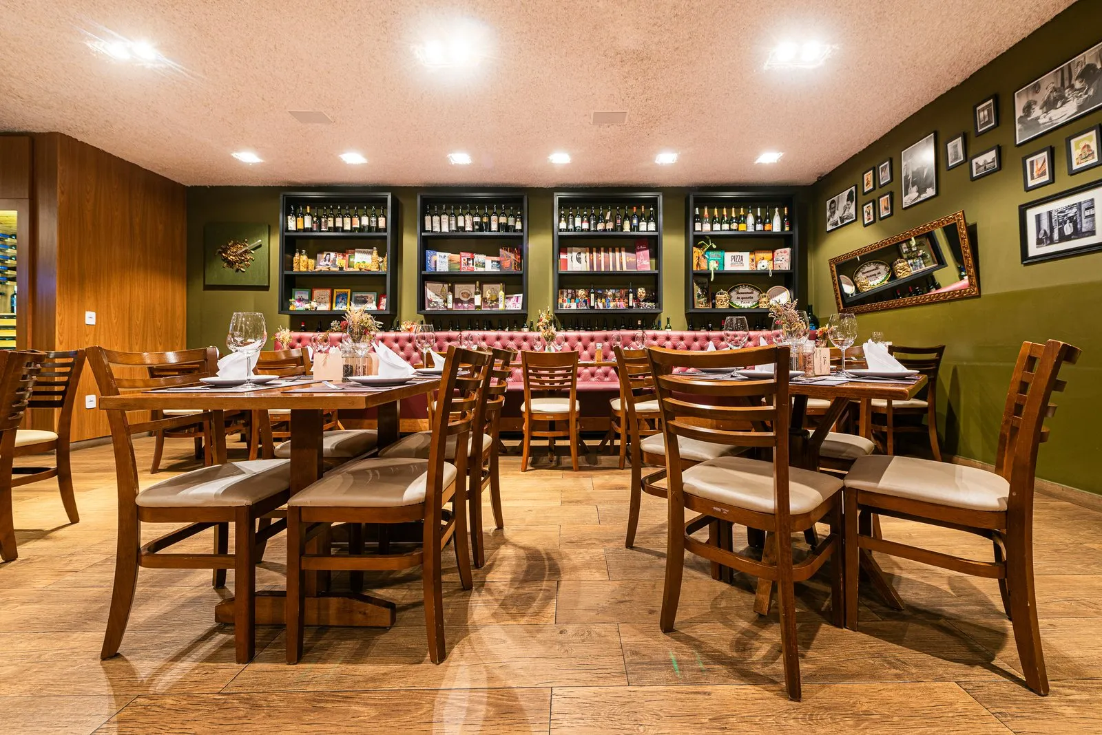 Fundo do salão da Cantina Castellammare com banco capitonê rosé, quatro nichos pretos com vinhos e livros, painel de madeira e mesas postas