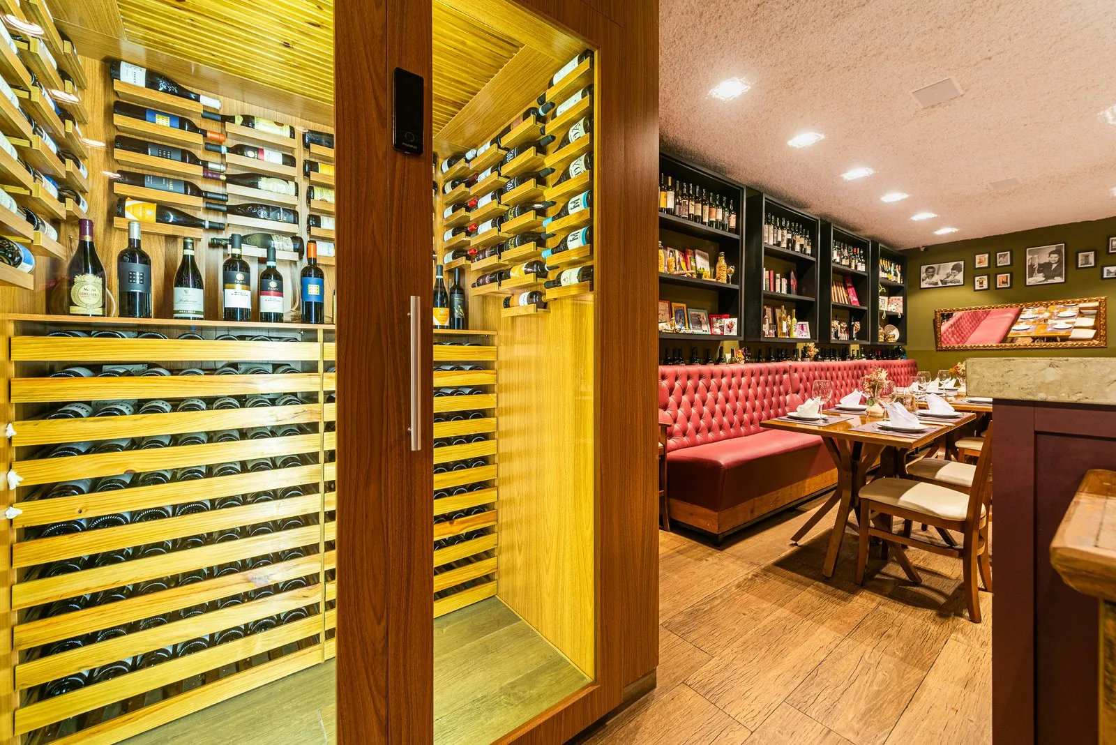 Adega de vinhos com prateleiras de madeira clara iluminadas na Cantina Castellammare, ao lado do salão com banco capitonê rosa e estantes pretas