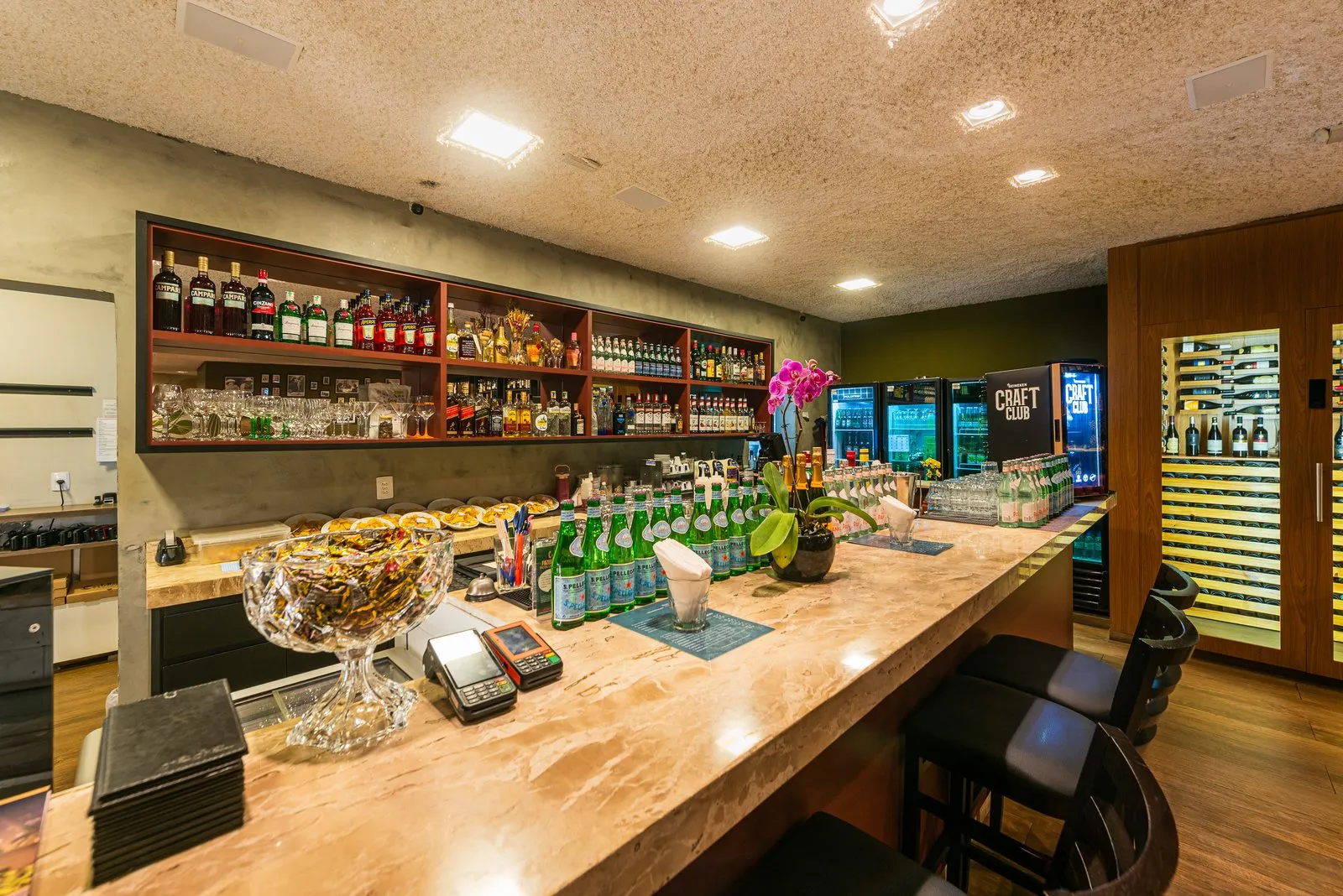 Bar da Cantina Castellammare com balcão em mármore, prateleira vermelha de destilados, orquídea, banquetas pretas e adega envidraçada ao fundo