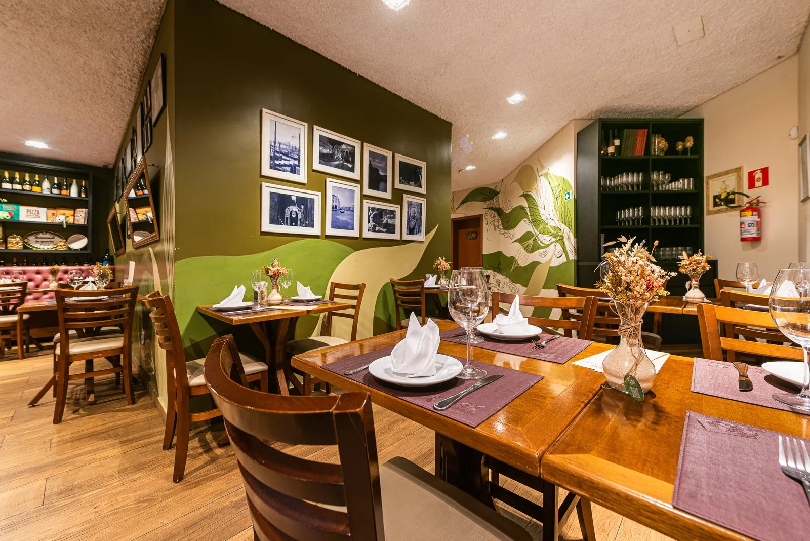 Salão da Cantina Castellammare com parede verde-oliva, fotos em preto e branco, mural de folhas e mesas de madeira com jogo americano lilás