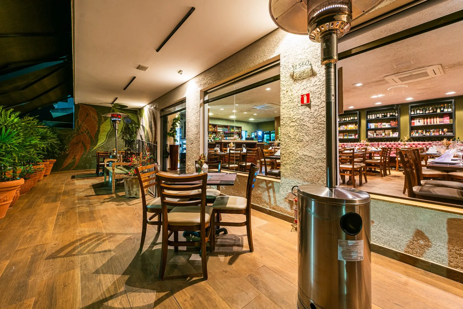 Varanda da Cantina Castellammare com aquecedor de inox, mesas de madeira e vasos de plantas, vista através das janelas para o salão iluminado