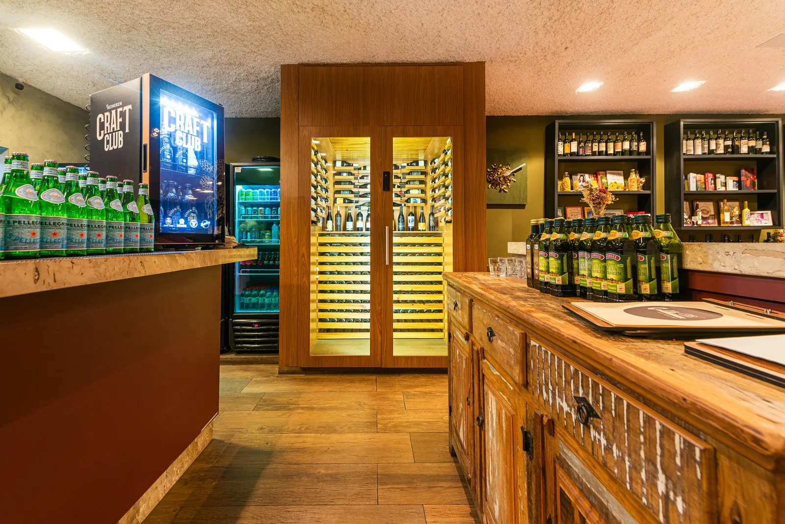 Adega climatizada da Cantina Castellammare com portas de vidro e prateleiras iluminadas, entre o balcão do bar e um aparador de madeira rústica