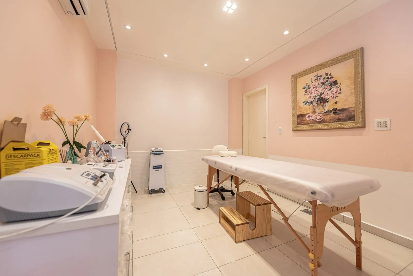 Sala com maca de madeira, equipamentos sobre a bancada e quadro de flores na parede
