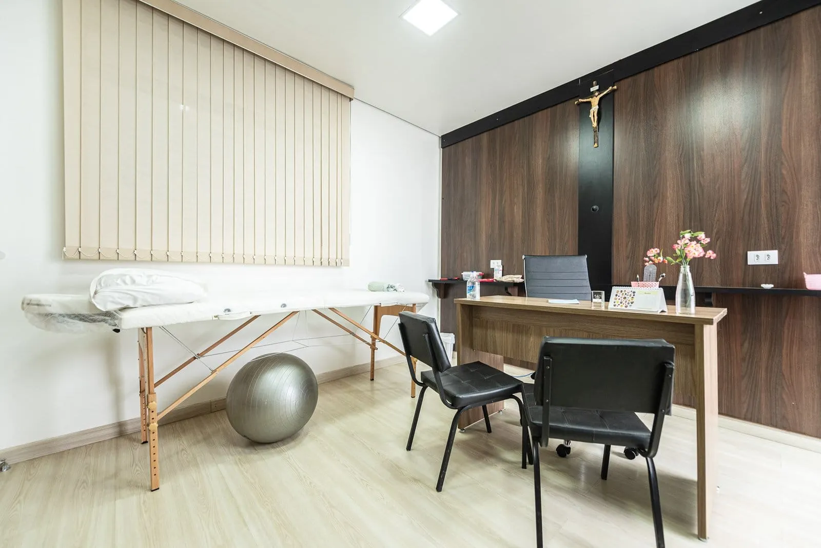 Consultório com mesa de madeira escura, maca, bola de pilates e persiana vertical