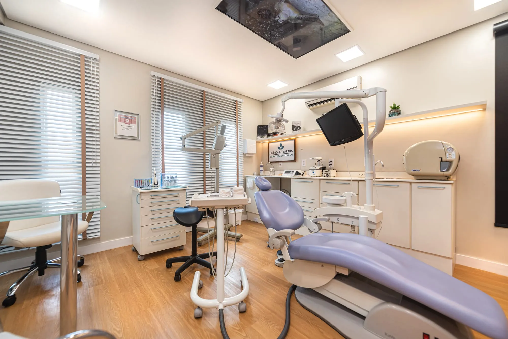 Consultório odontológico com cadeira de estofado lilás em primeiro plano, bancadas brancas, monitor em braço articulado, mesa de vidro e o painel iluminado com imagem de céu e mar no teto
