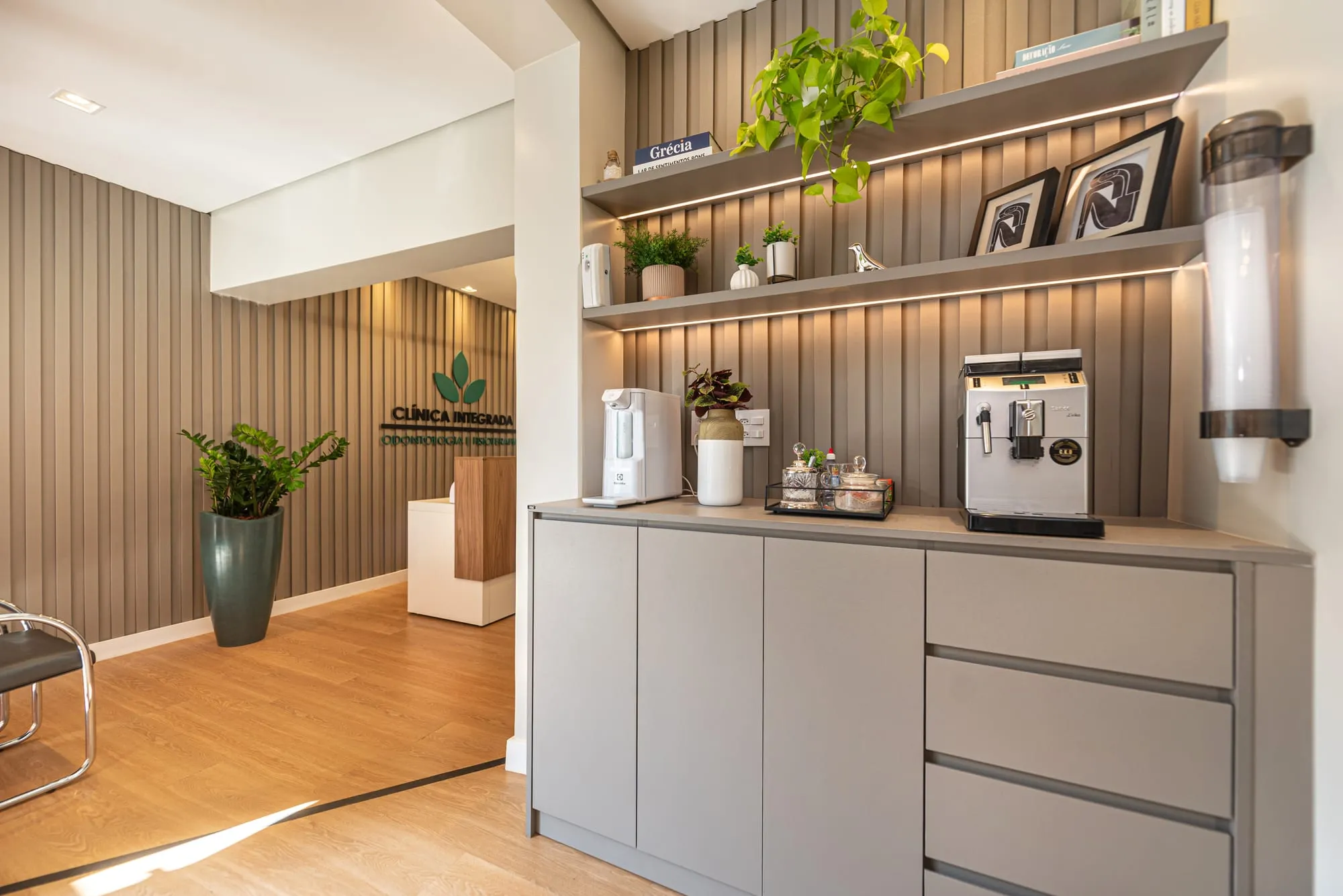 Espaço café com armário cinza-claro, máquina de café expresso, purificador de água e prateleiras iluminadas com plantas e quadros sobre o painel de ripas