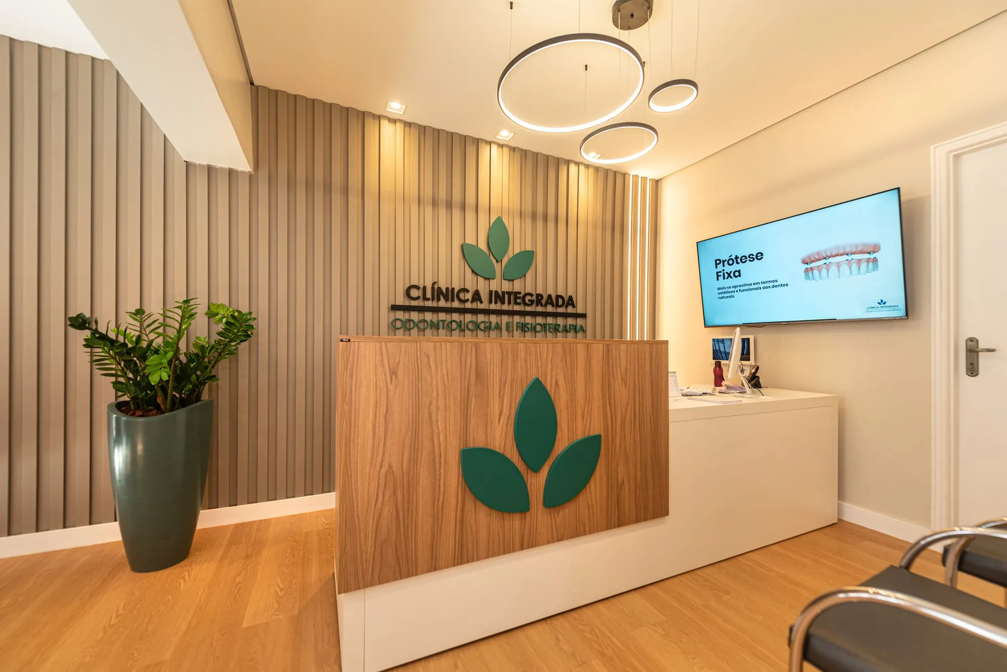 Recepção da Clínica Integrada, com o balcão de madeira e tampo branco com três folhas verdes na frente, a parede de ripas com o logotipo em relevo, uma luminária de três anéis de LED e um vaso alto com planta