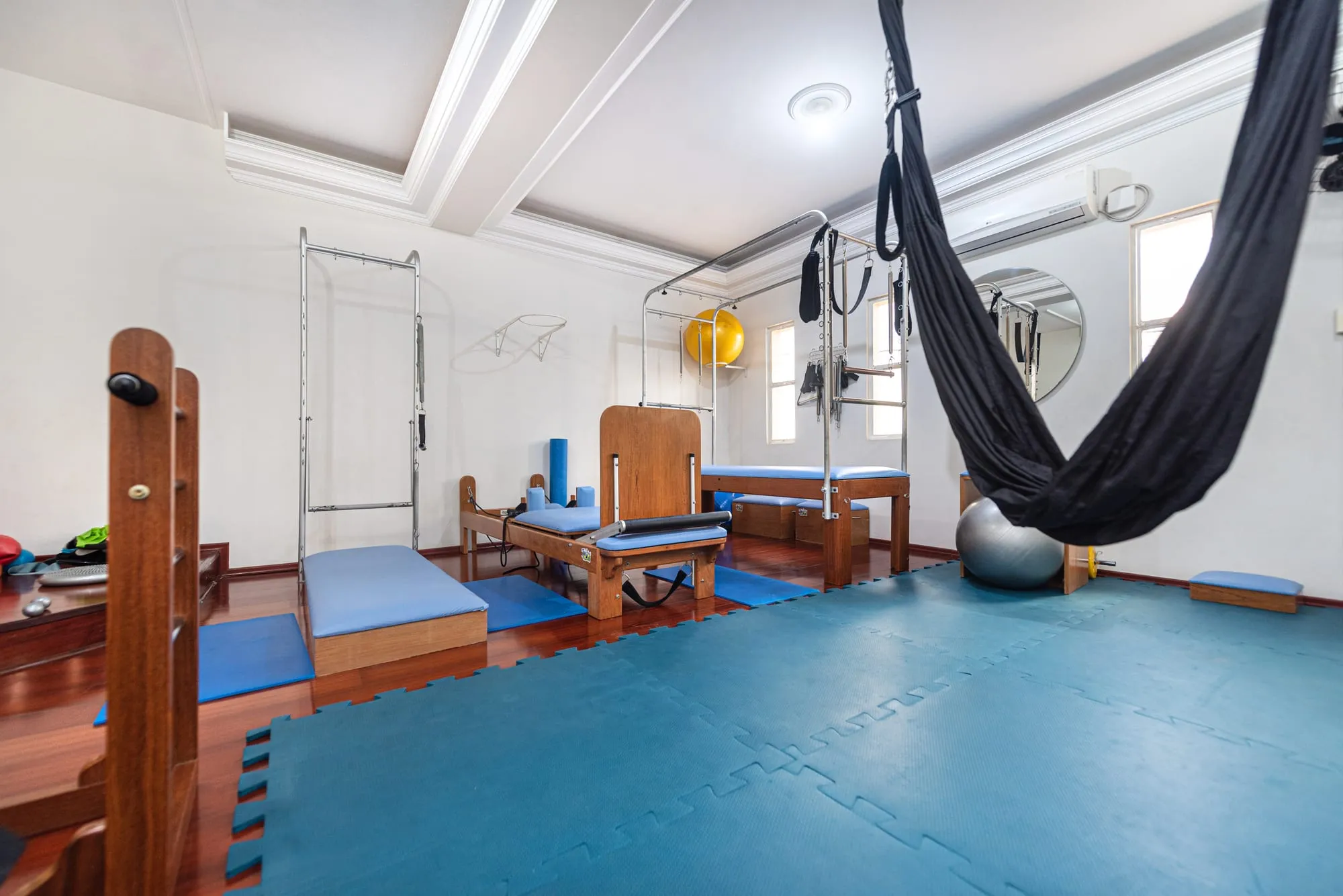 Studio de Pilates com piso emborrachado azul-petróleo, aparelhos de madeira com estofado azul, a estrutura metálica do trapézio e uma rede preta pendurada