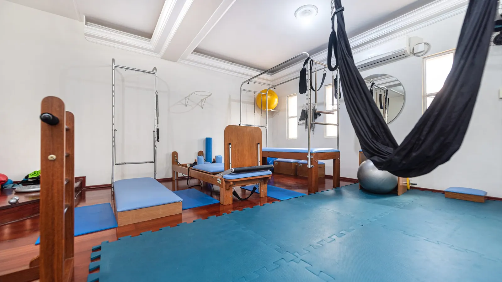 Studio de Pilates da Clínica Integrada, com piso emborrachado azul-petróleo, aparelhos de madeira com estofado azul, a estrutura metálica do trapézio e uma rede preta pendurada
