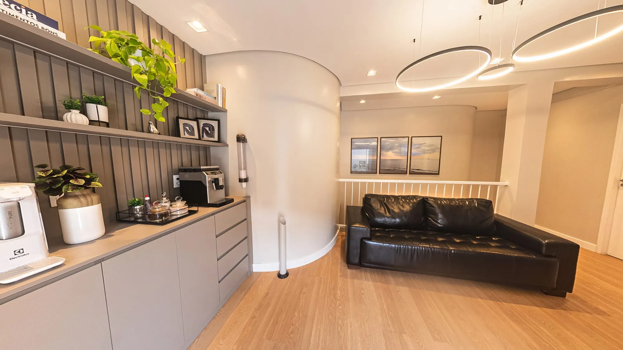 Sala de espera com sofá de couro preto diante de três quadros de paisagens de mar, a parede curva branca, a luminária de anéis no teto e o espaço café à esquerda