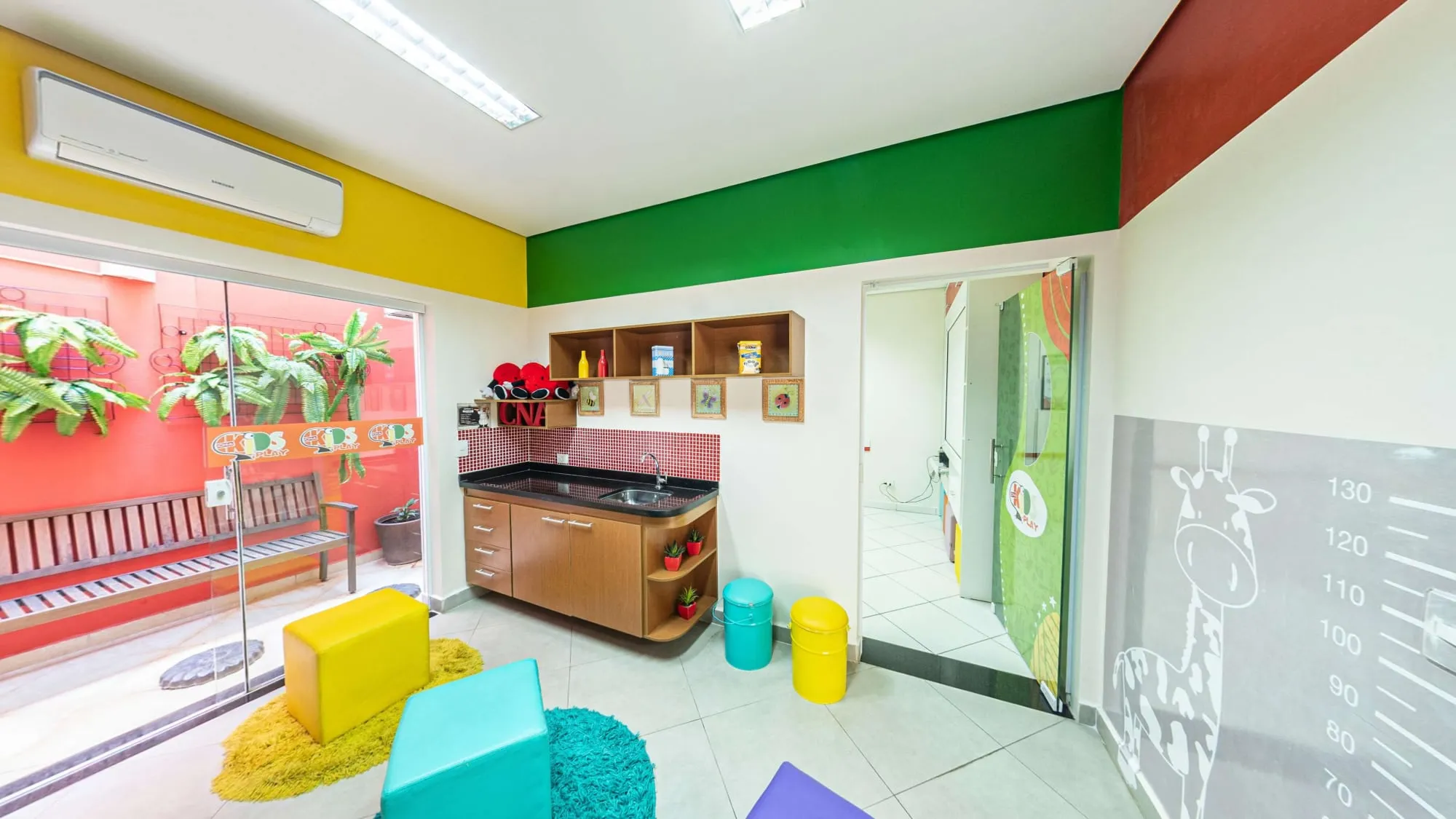 Sala Kids Play do CNA Itapetininga, com paredes amarela, verde e vermelha, pia baixa, puffs coloridos e régua de altura com girafa