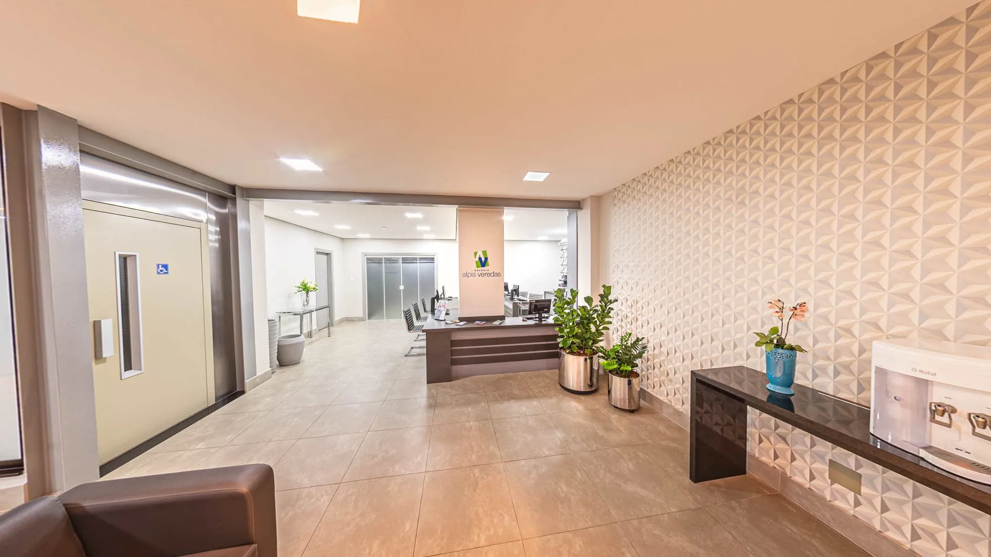 Área de espera do Colégio Alpis Veredas, com sofá de couro marrom, parede de revestimento 3D branco, vasos de plantas e o balcão de atendimento com o logotipo ao fundo