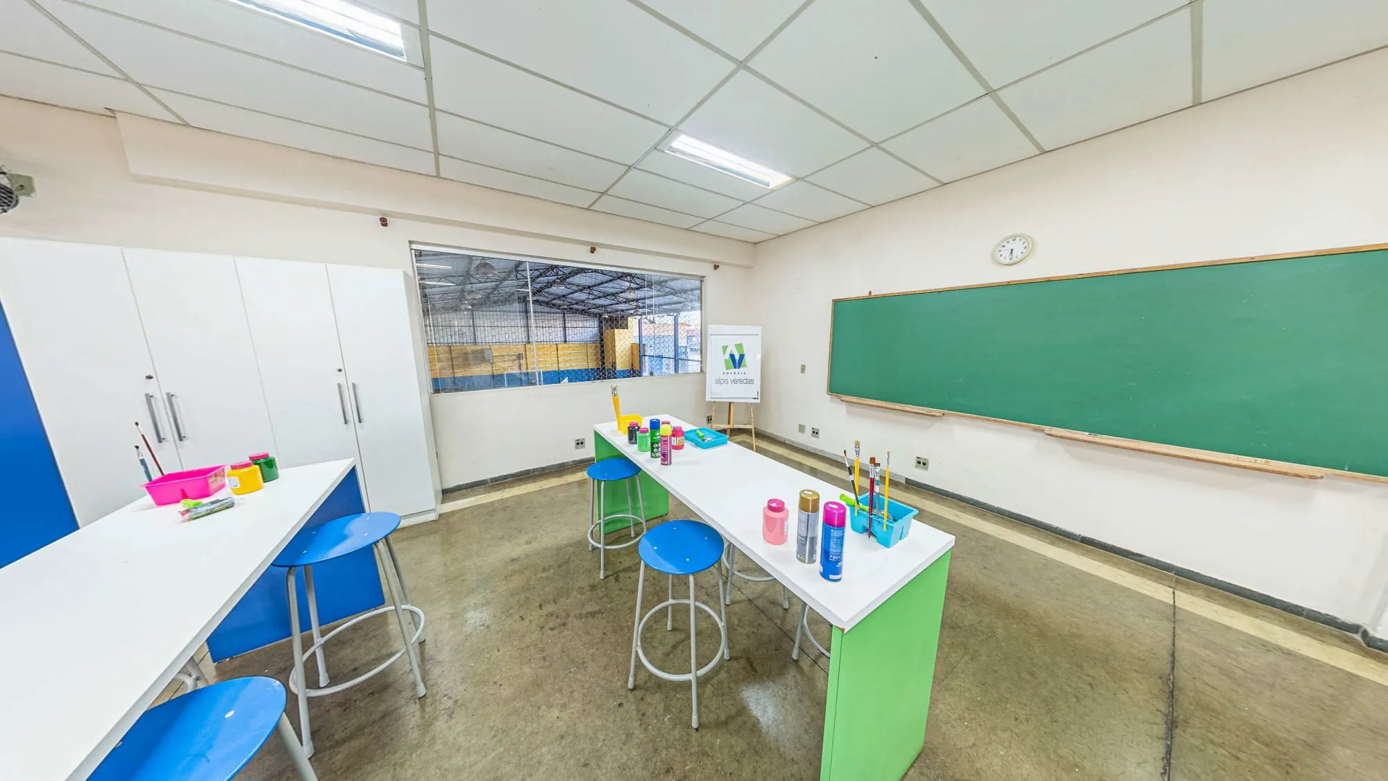 Laboratório de artes do Colégio Alpis Veredas, com bancadas brancas e verdes, banquetas azuis, tintas e pincéis, lousa verde e janela para a quadra