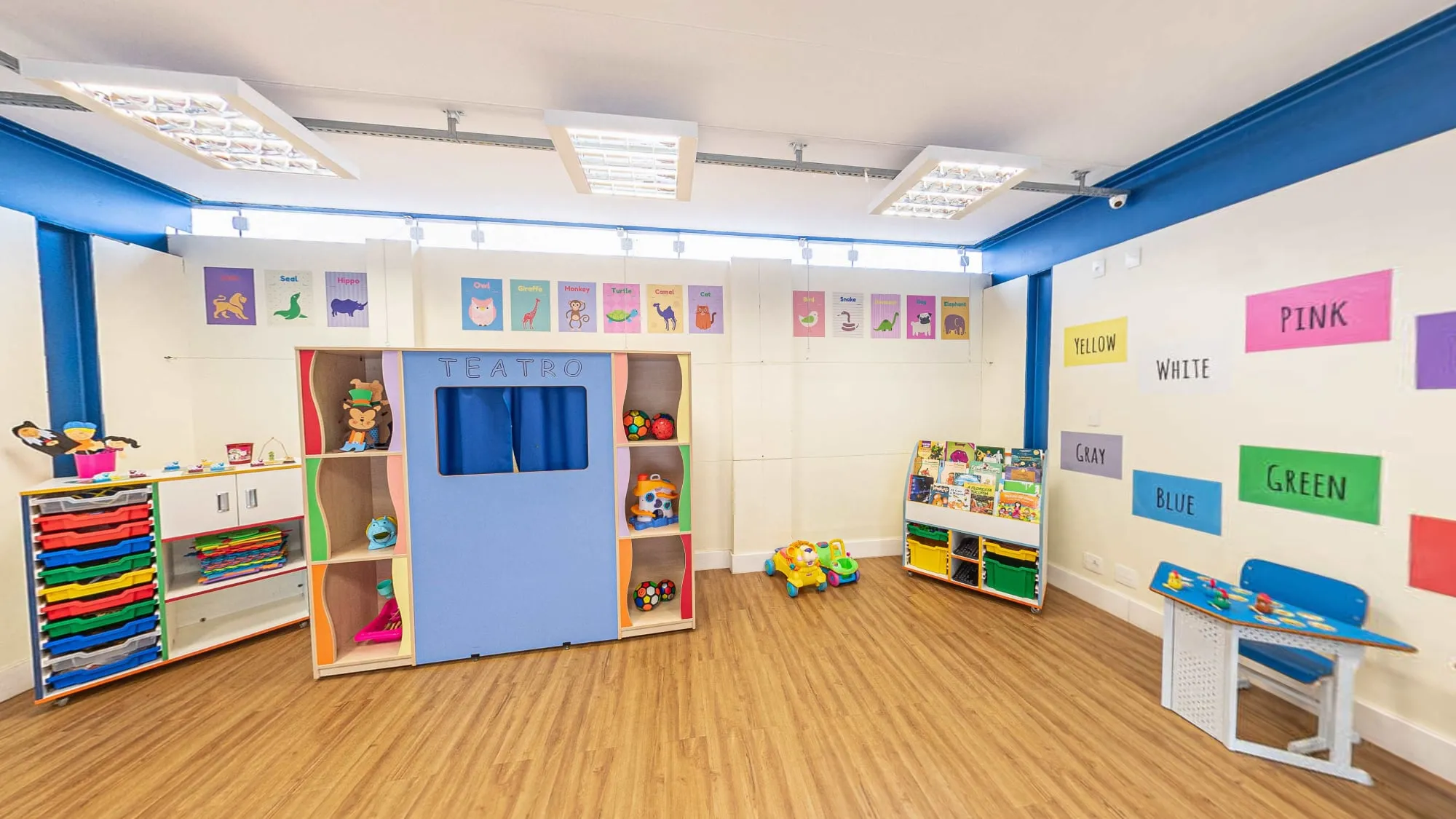 Sala de atividades do Colégio Alpis Veredas, com teatrinho de fantoches azul, estantes de brinquedos, mesinhas coloridas e cartazes de cores em inglês