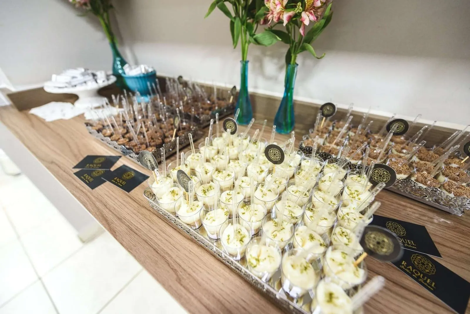 Mesa de doces em copinhos, com vasos de flores, servida na reinauguração de um salão de beleza