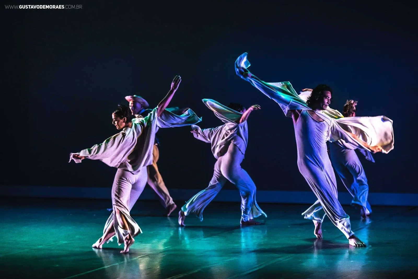 Bailarinos de figurino branco em movimento no palco, sob luz azul e verde