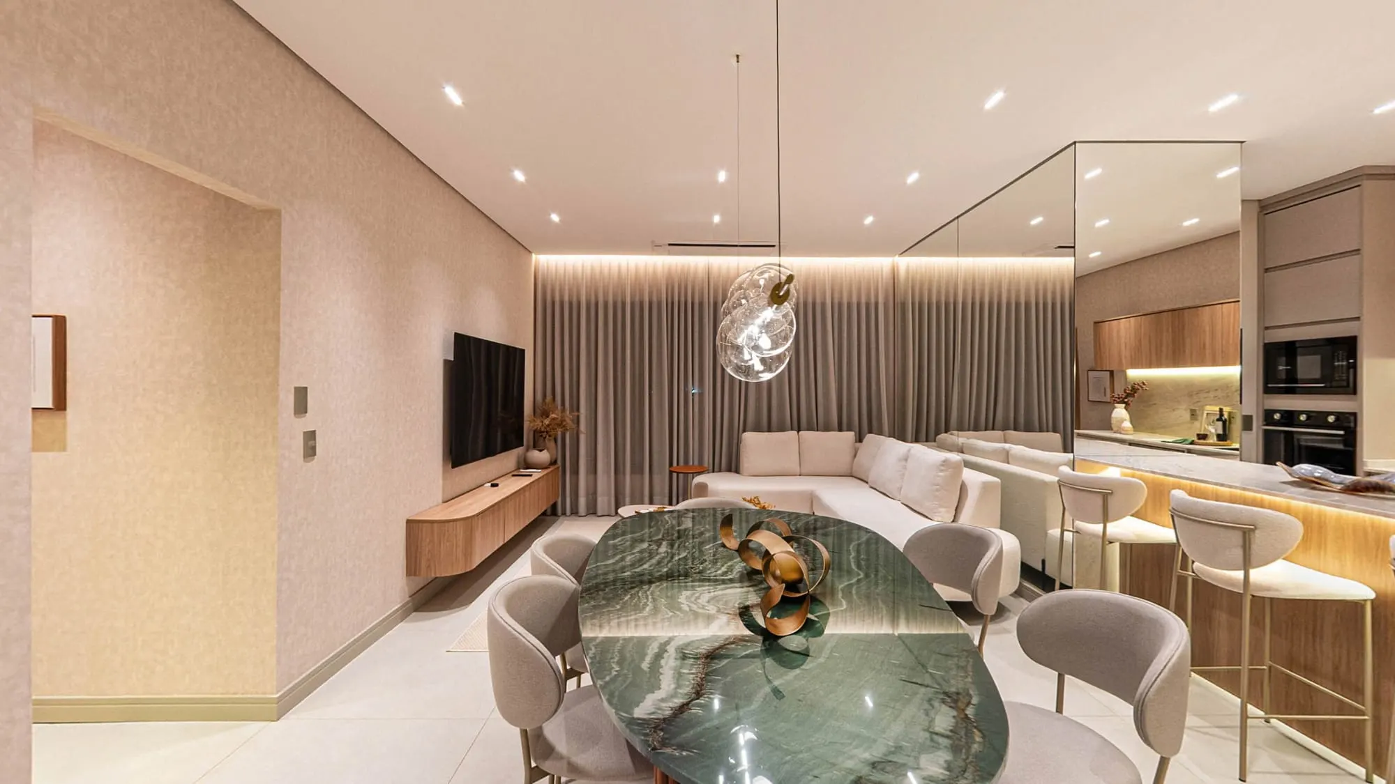 Sala de jantar do decorado do Grand’Oro Vila Barth, com mesa oval de mármore verde e cadeiras claras, o sofá e a TV da sala de estar ao fundo e a ilha da cozinha com banquetas à direita