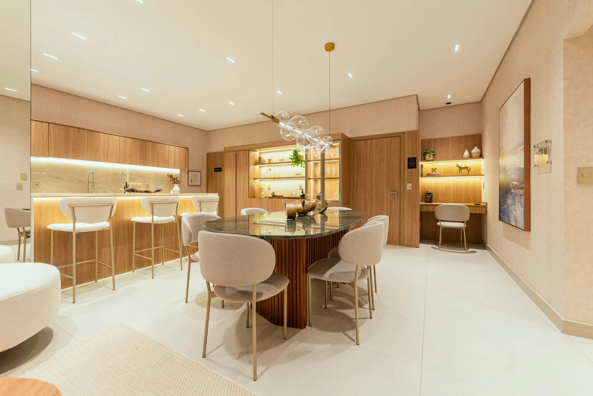 Sala de jantar do apartamento decorado do Grand'Oro Vila Barth, com mesa oval de mármore verde, lustre de bolhas de vidro, cristaleira iluminada e a bancada da cozinha ao fundo