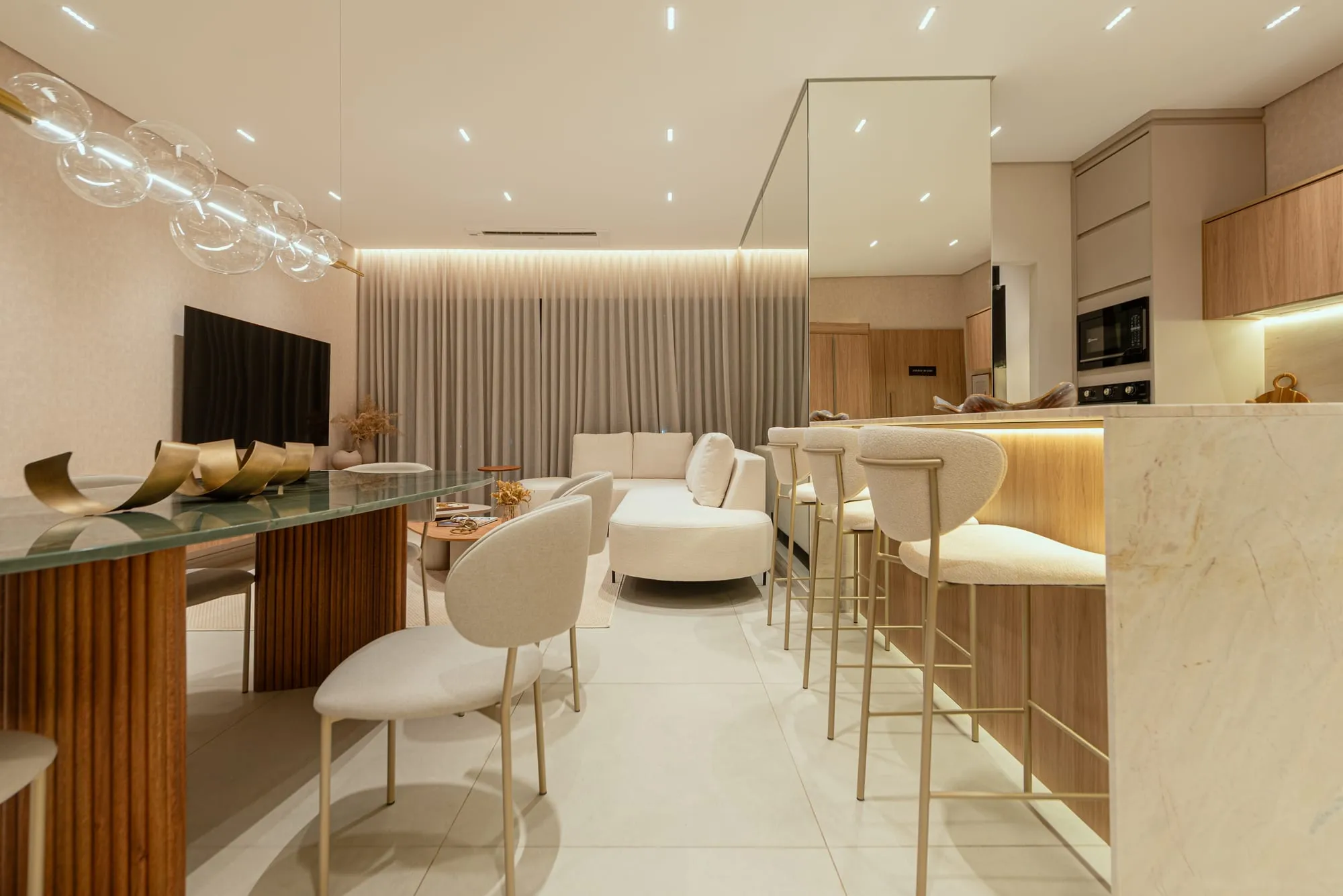 Living integrado visto da mesa de jantar: sofá curvo e TV na sala de estar e a ilha de pedra com banquetas à direita